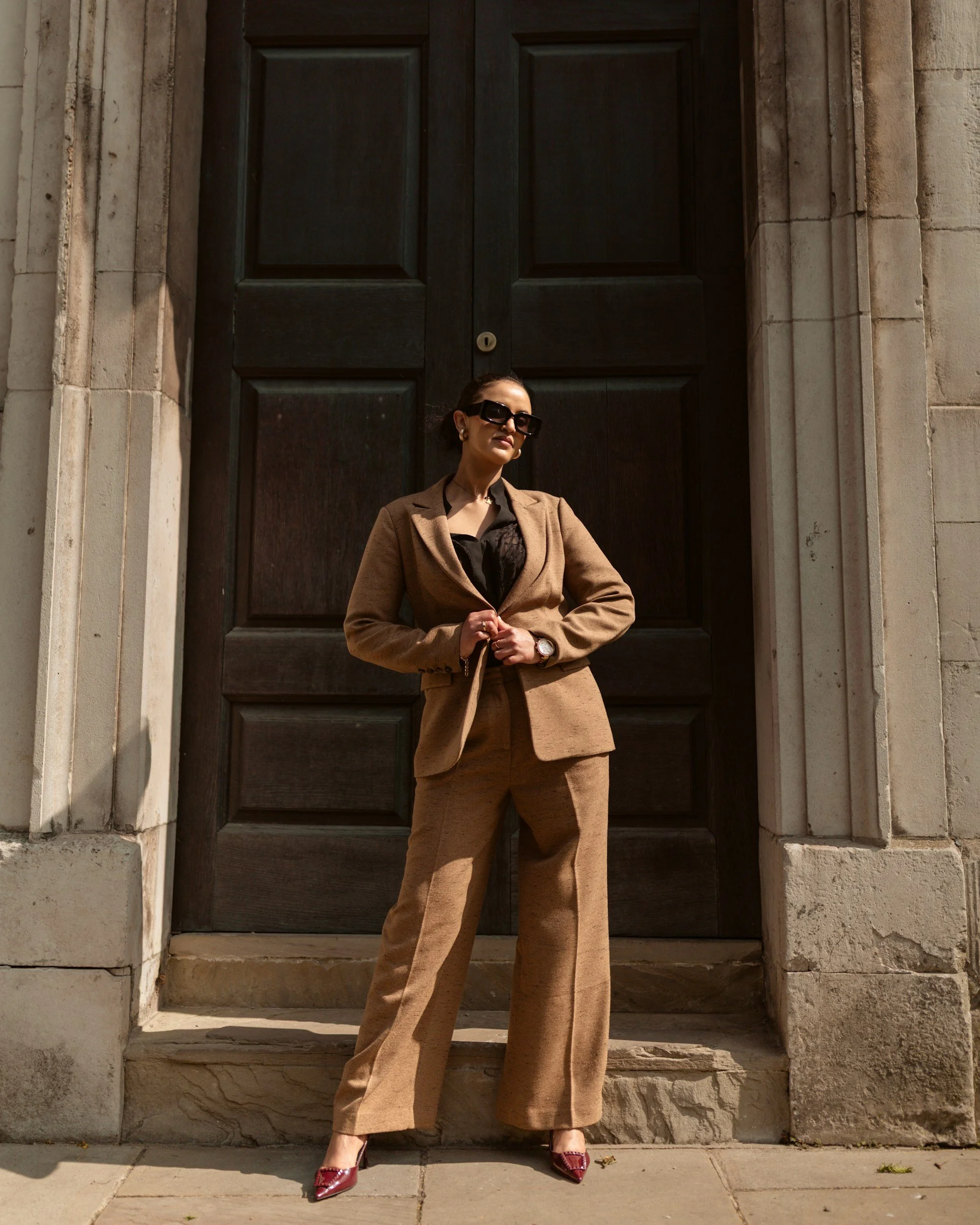 A woman dressed in a brown suit, black blouse, and red heels standing in front of a large dark wooden door with stone walls on either side.