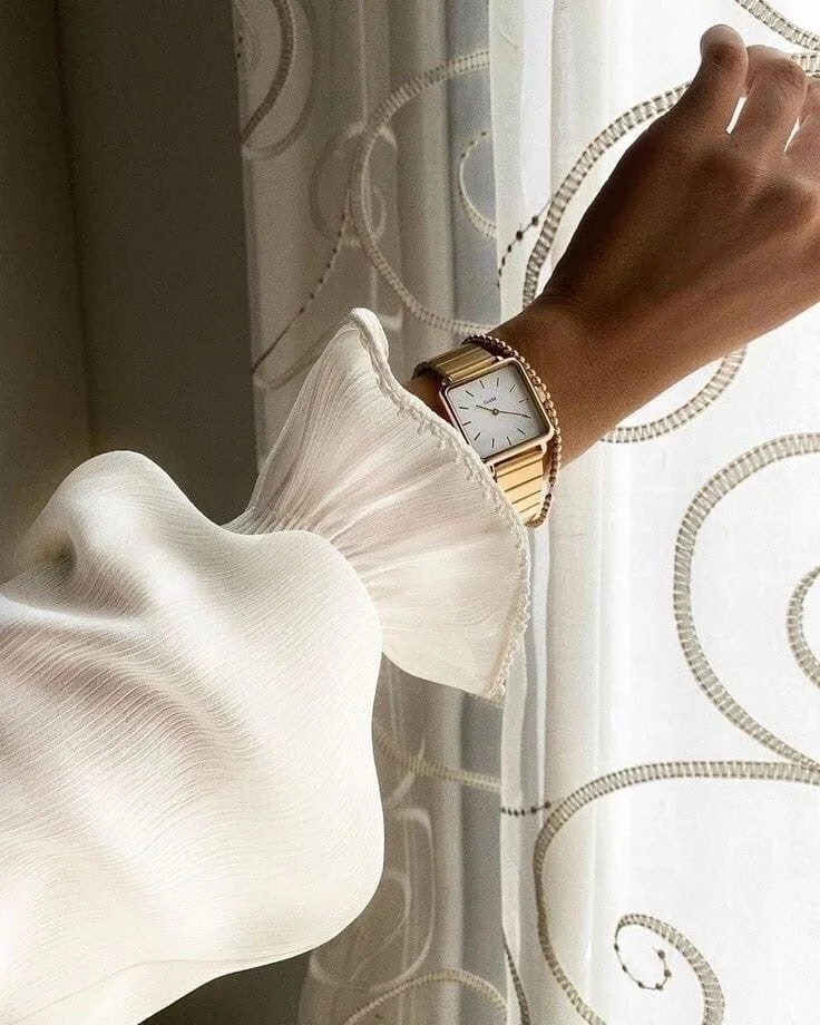 A person's arm extends towards a window, showing a gold wristwatch with a rectangular face and white dial, wearing a pearl bracelet. The person is dressed in a white, long-sleeved blouse with a ruffled cuff, and sheer fabric. The background includes a sheer curtain with swirling line patterns.