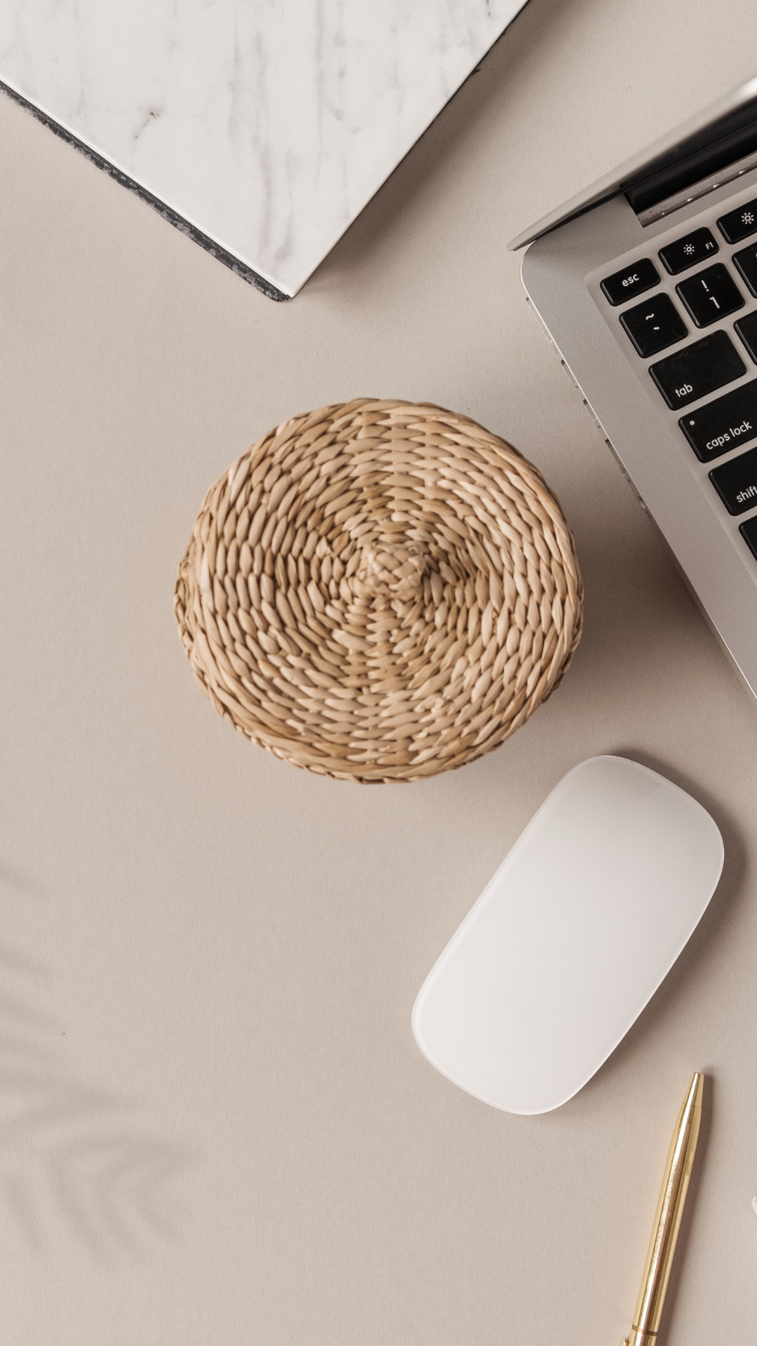 Top view of a workspace with a woven basket, a white mouse, a gold pen, a laptop, and a marble-patterned notebook on a beige surface.