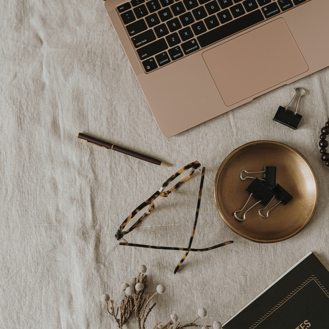 Flat lay of a workspace with a laptop, a pair of tortoise shell glasses, a black pen, a golden plate with black binder clips, a black notebook, and a dried flower branch on a beige fabric surface.