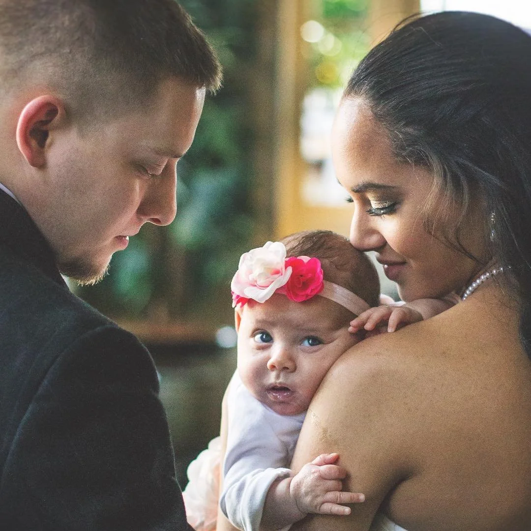 A young couple holding their newborn baby girl, who is wearing a pink headband with flowers, close to their faces with their eyes closed, creating an intimate moment.