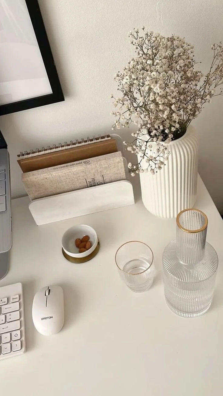 A white desk with a computer monitor, keyboard, and a white mouse. A white vase with white baby's breath flowers, a small white dish with almonds, and two clear glass vases with gold accents.