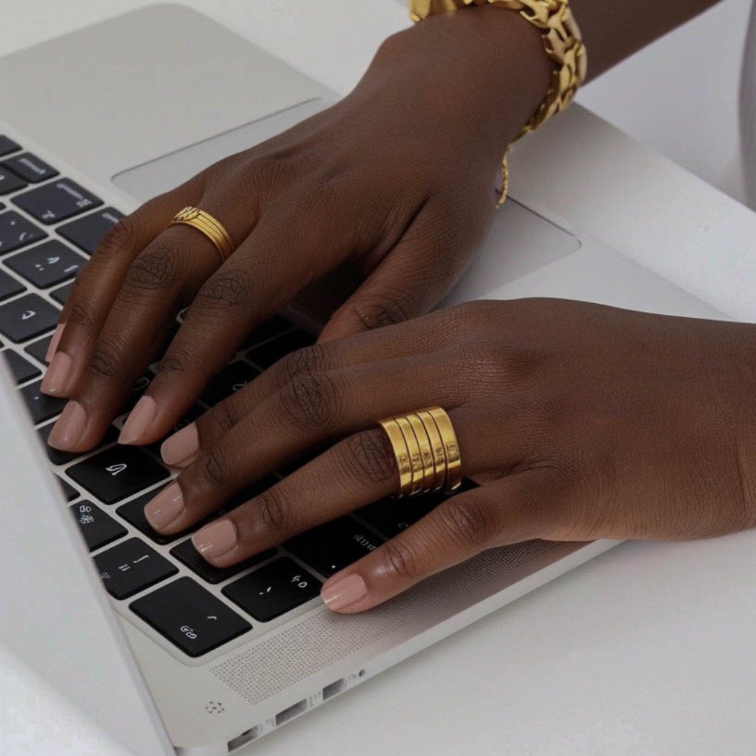 Close-up of dark-skinned hands with gold rings and bracelets typing on a silver laptop keyboard.