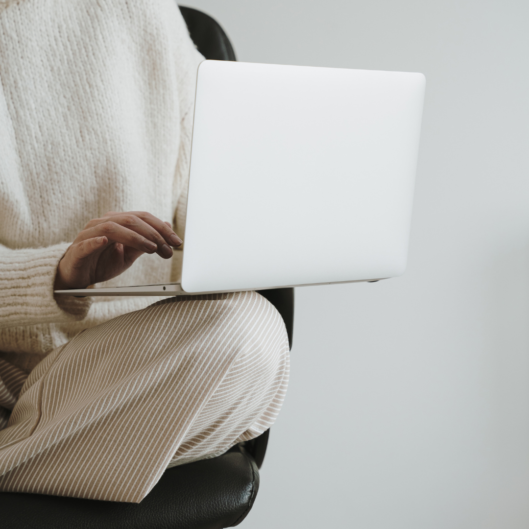 Person sitting in a chair using a silver laptop, wearing beige striped pants and a cream-colored knit sweater.