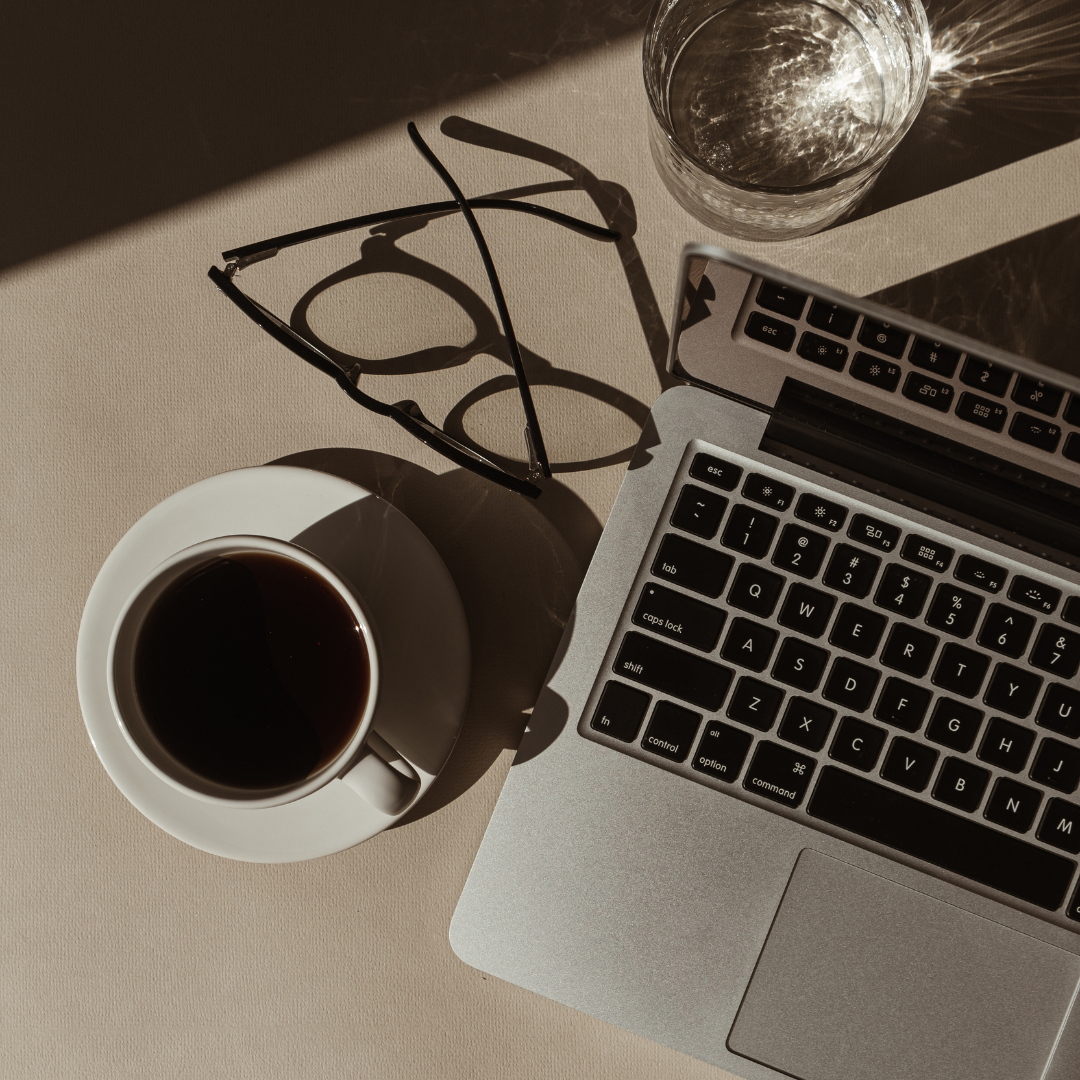 A laptop, a cup of coffee on a saucer, a pair of eyeglasses, and a glass of water on a beige surface with shadows.