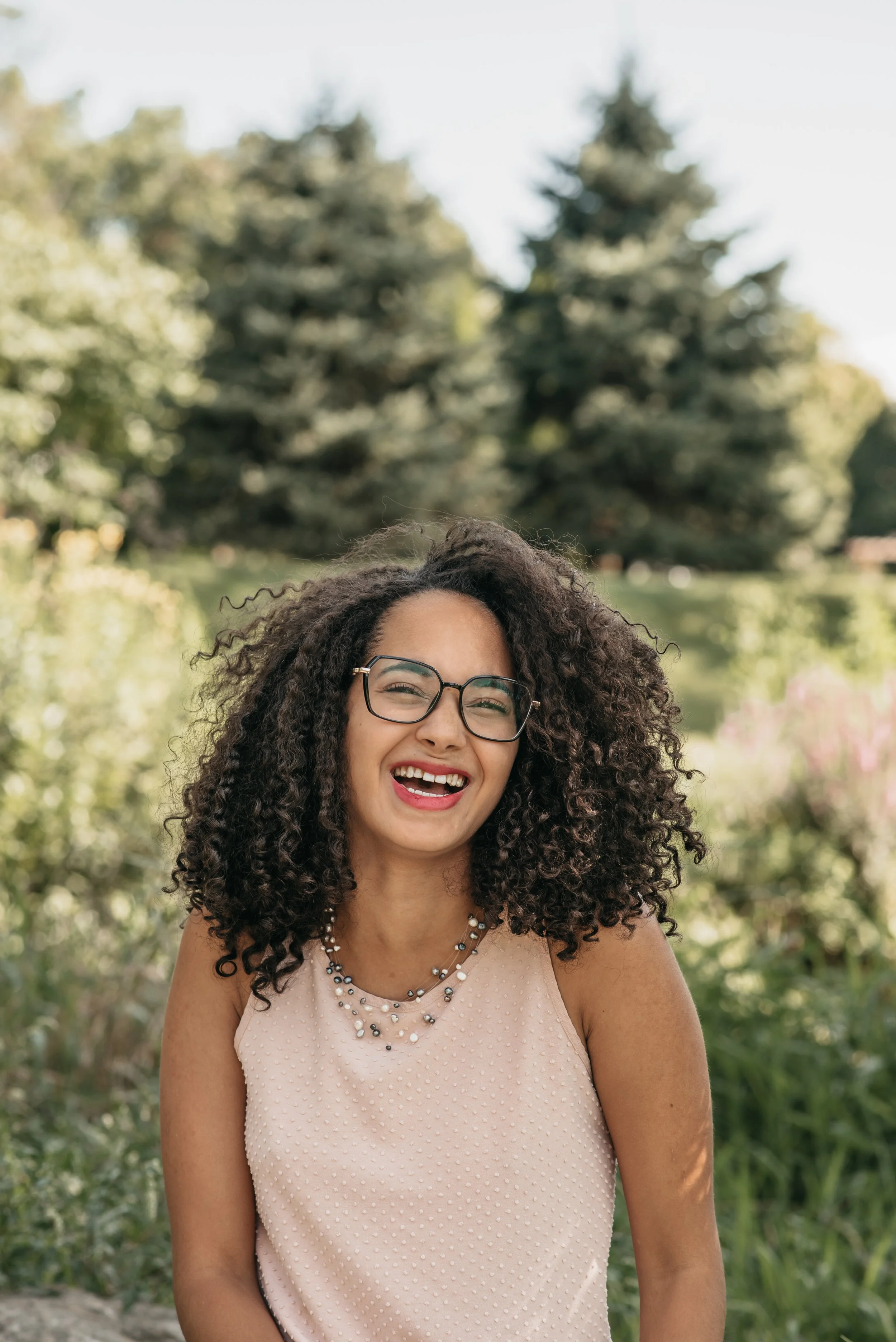 A woman with curly dark hair wearing glasses, a light-colored sleeveless top, and layered necklaces, smiling outdoors with greenery and trees in the background.