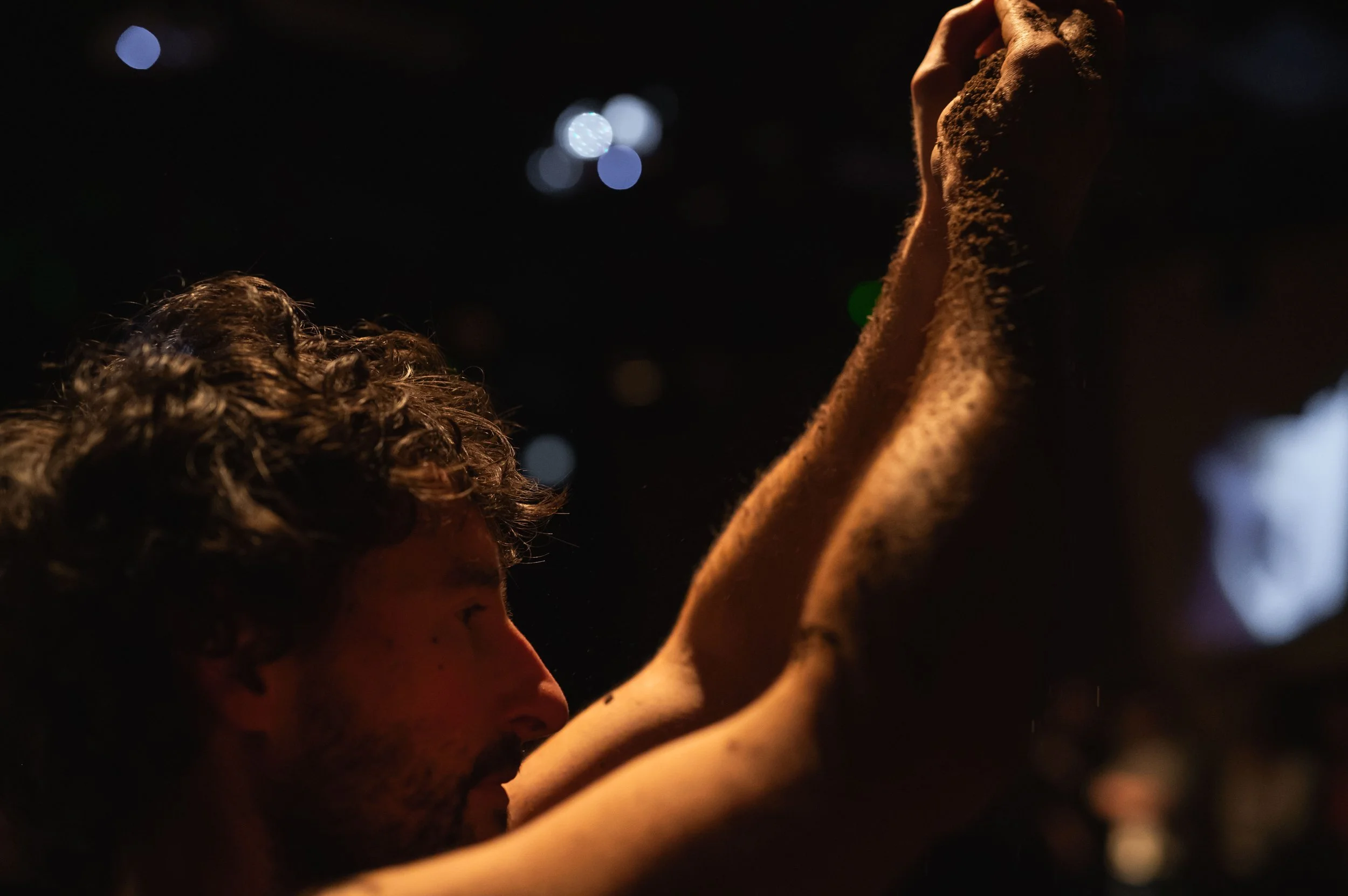 Un homme avec des cheveux bouclés, vu de profil, tenant une pièce de tissu ou une pièce de vêtement dans ses mains dans un environnement sombre avec des lumières floues en arrière-plan.