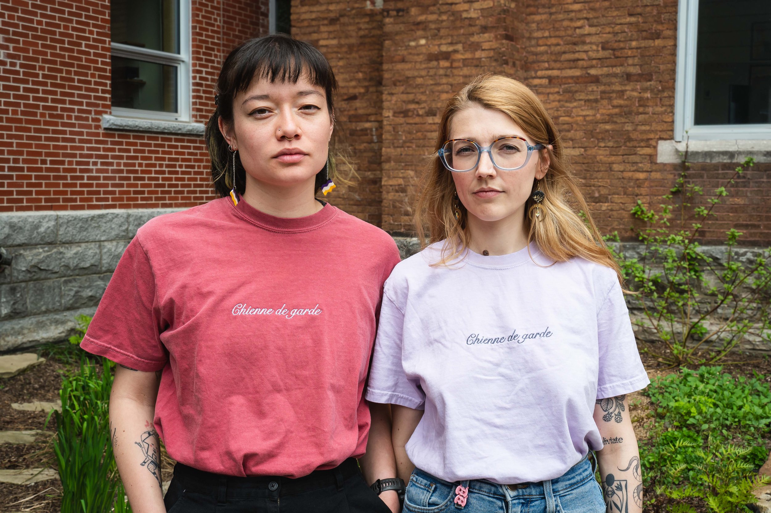 Deux jeunes femmes debout devant un mur de briques et un bâtiment, portant des t-shirts avec l'inscription 'Chienne de garde', l'une en rouge et l'autre en violet, avec des tatouages et des boucles d'oreilles colorées.