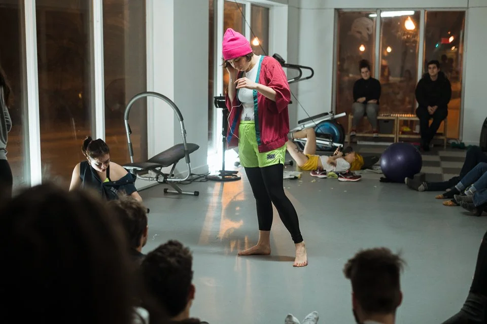 Jeune femme en tenue décontractée, portant un bonnet rose, acceptant une bouteille d'eau dans une salle en intérieur, avec des groupes de personnes qui assistaient à une activité ou un cours.