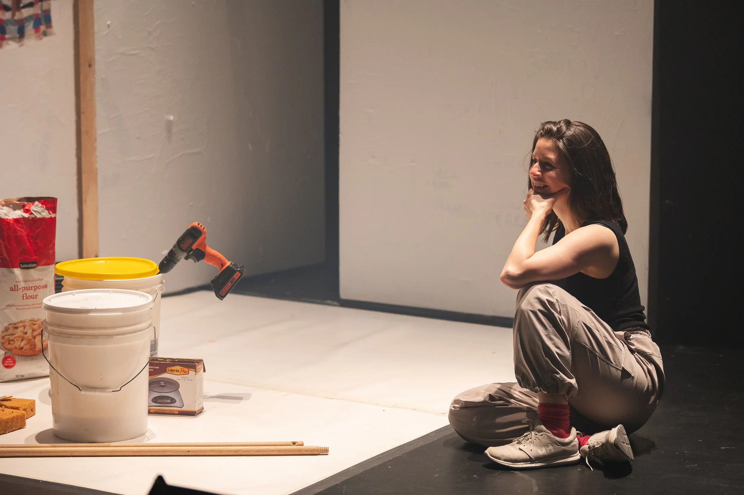 Une femme assise par terre dans un studio de création, entourée de matériaux de construction et d'outils, souriant et en train de réfléchir.