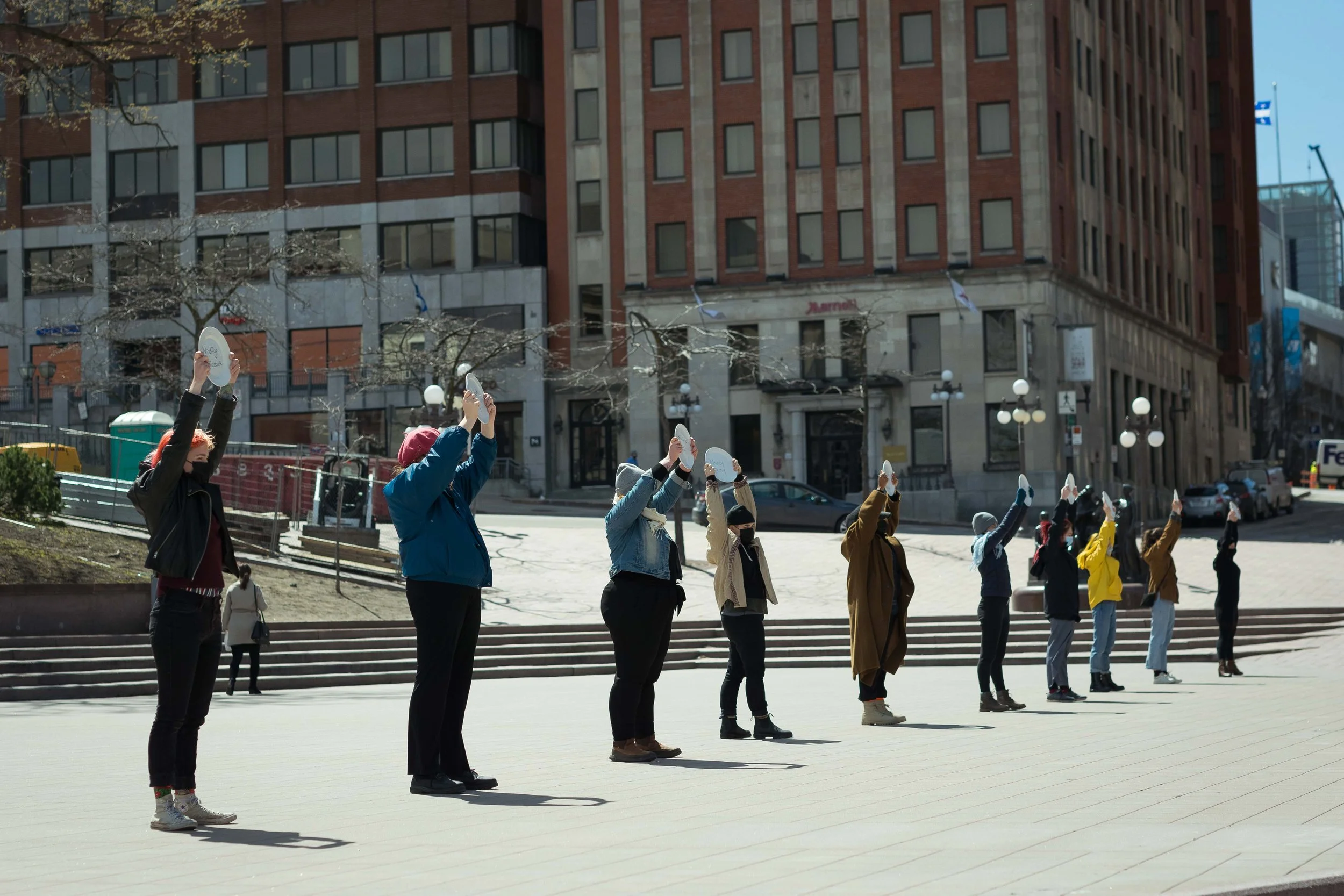 Groupe de personnes portant des masques faciaux dans une file ordonnée à l'extérieur devant un bâtiment en milieu urbain, probablement lors d'une performance artistique du théâtre de l'Impie