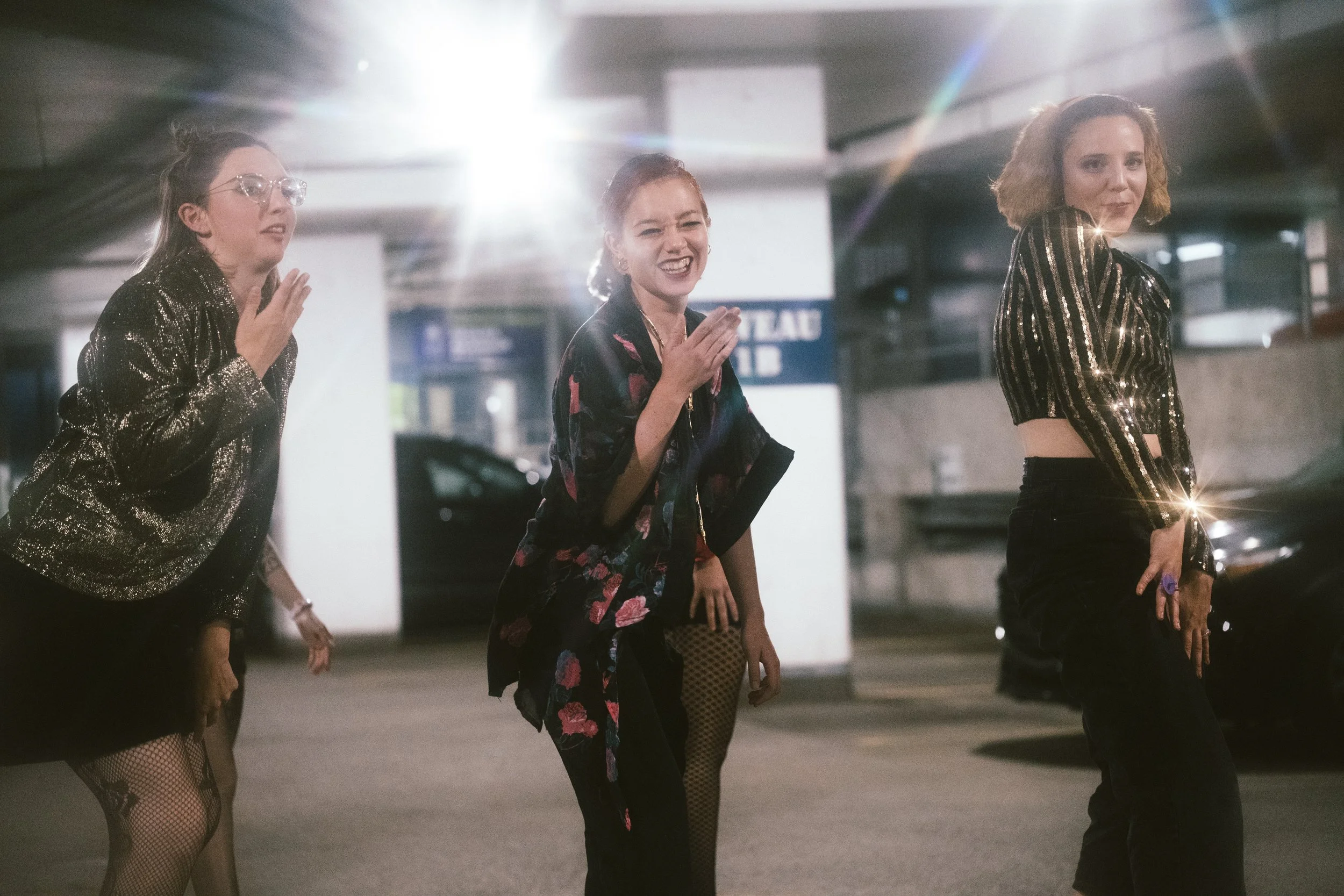 Trois femmes dans une scène urbaine la nuit, vêtues de vêtements brillants, souriantes et enjouées.