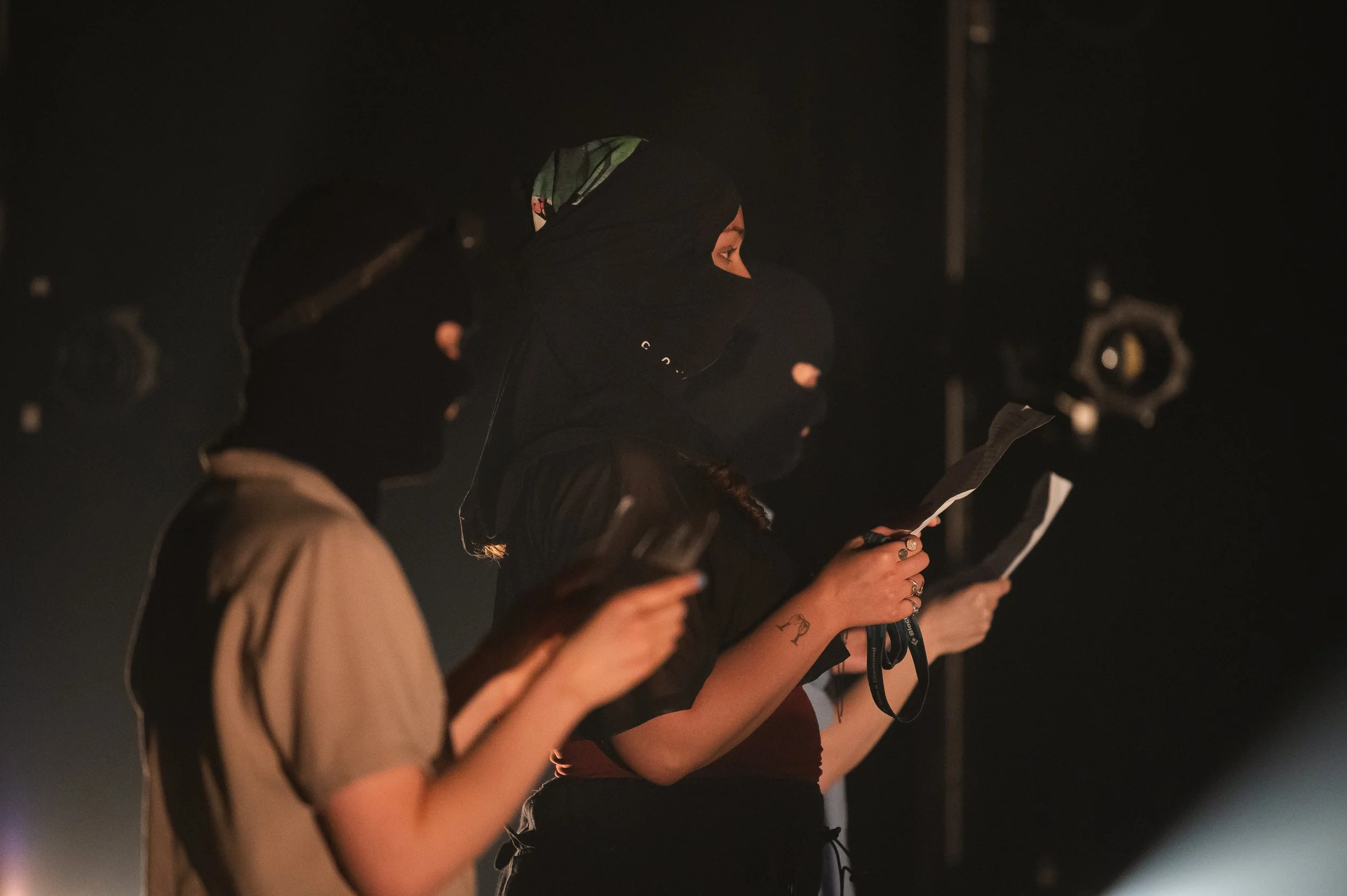 Trois femmes portant des cagoules s noirs se tiennent sur scène avec  des feuilles de papier, lors d'une performance. 