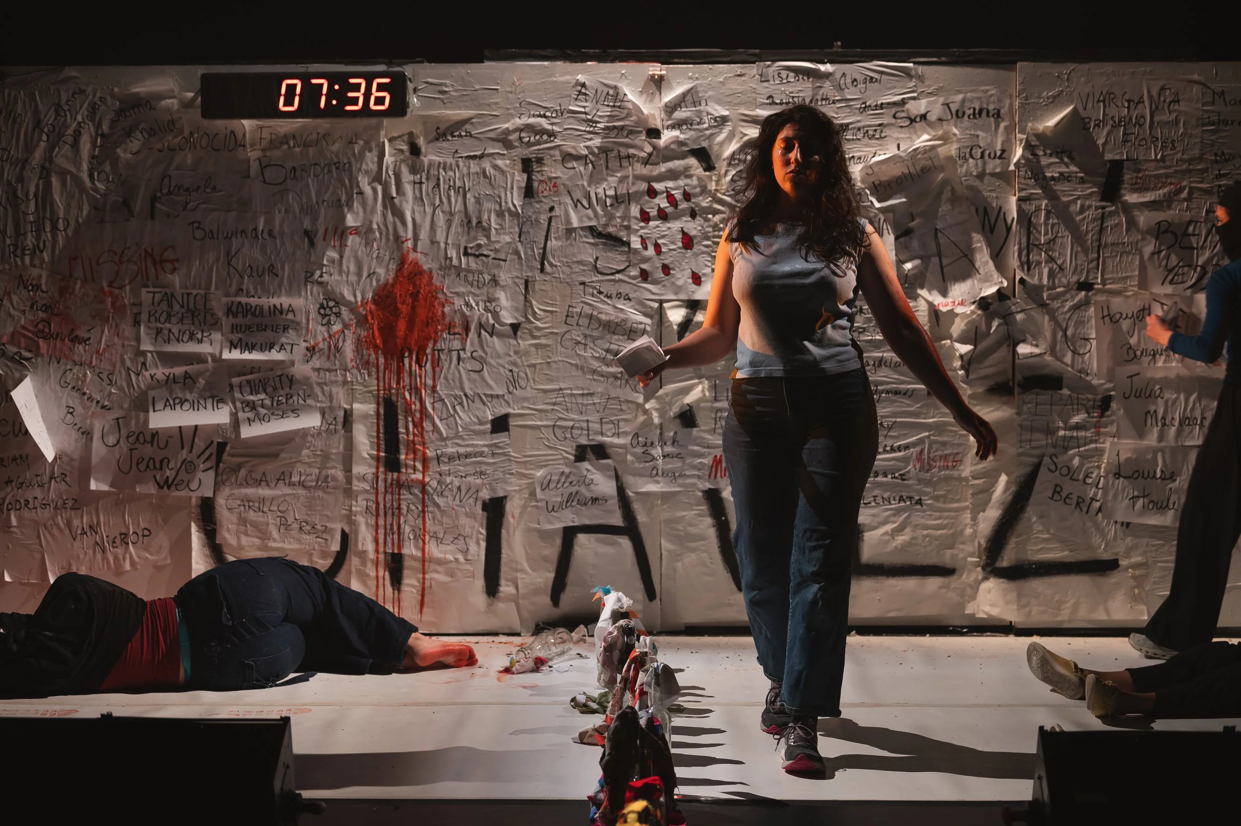 Une scène de pièce de théâtre ou performance artistique avec une femme au centre qui marche, tenant un objet dans la main. En arrière-plan, un mur de papier avec du texte écrit, des signatures et un dessin de gouttes de sang. Sur le sol, une personne