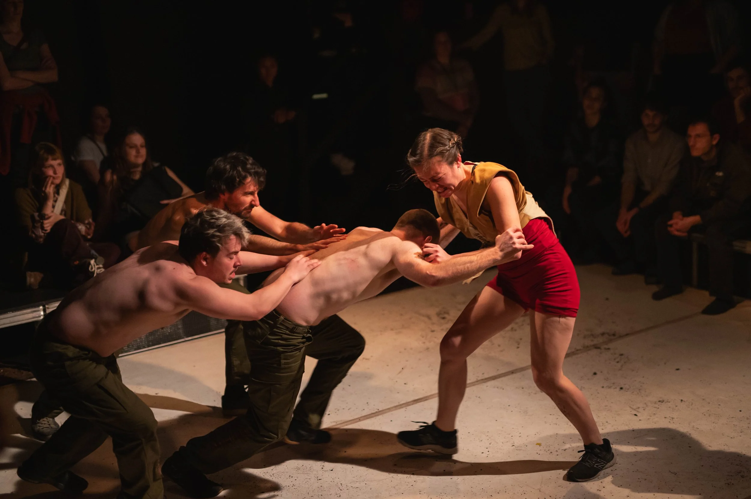 Une performance théâtrale représentant une scène de combat intense avec plusieurs acteurs, certains nus, avec un public regardant en arrière-plan.