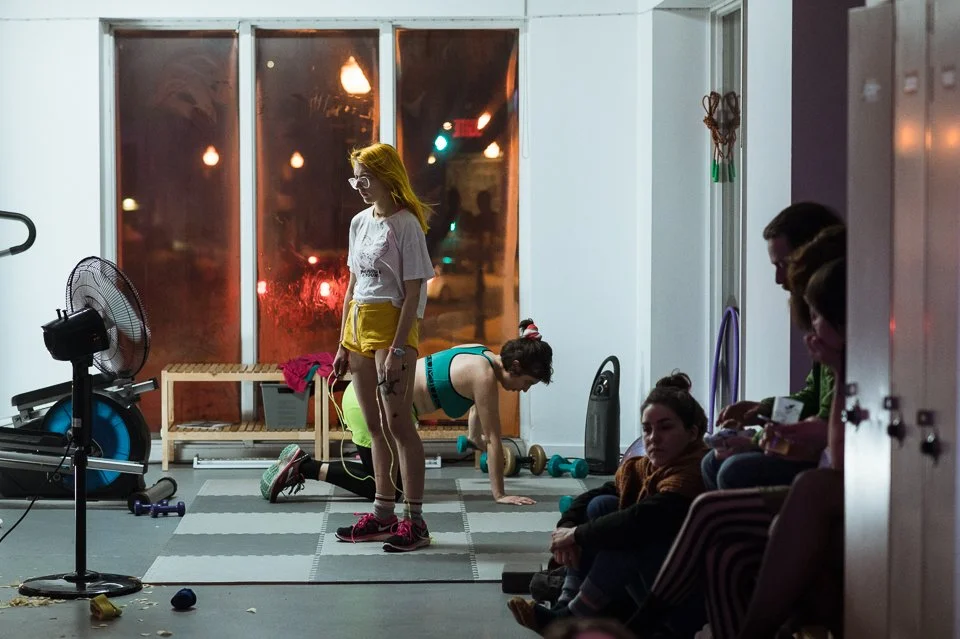 Groupe de personnes dans une salle de sport ou de fitness, avec une femme en train de faire des exercices au sol, un homme et une femme entre autres, dans une ambiance intérieure avec des fenêtres en arrière-plan