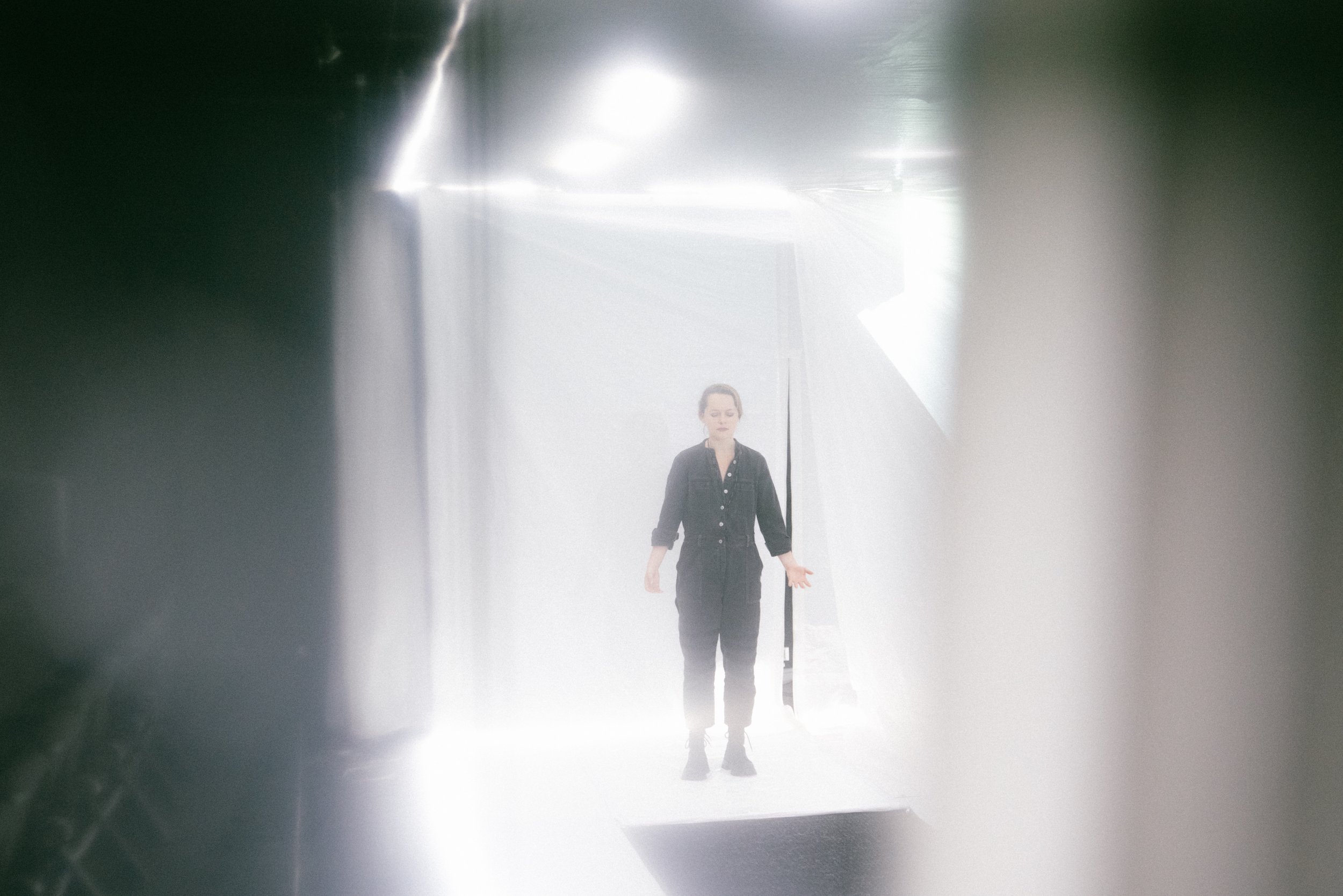 Une femme en vêtements noirs se tient au centre, dans un espace lumineux et flou, avec des murs blanc et des lumières vives autour.