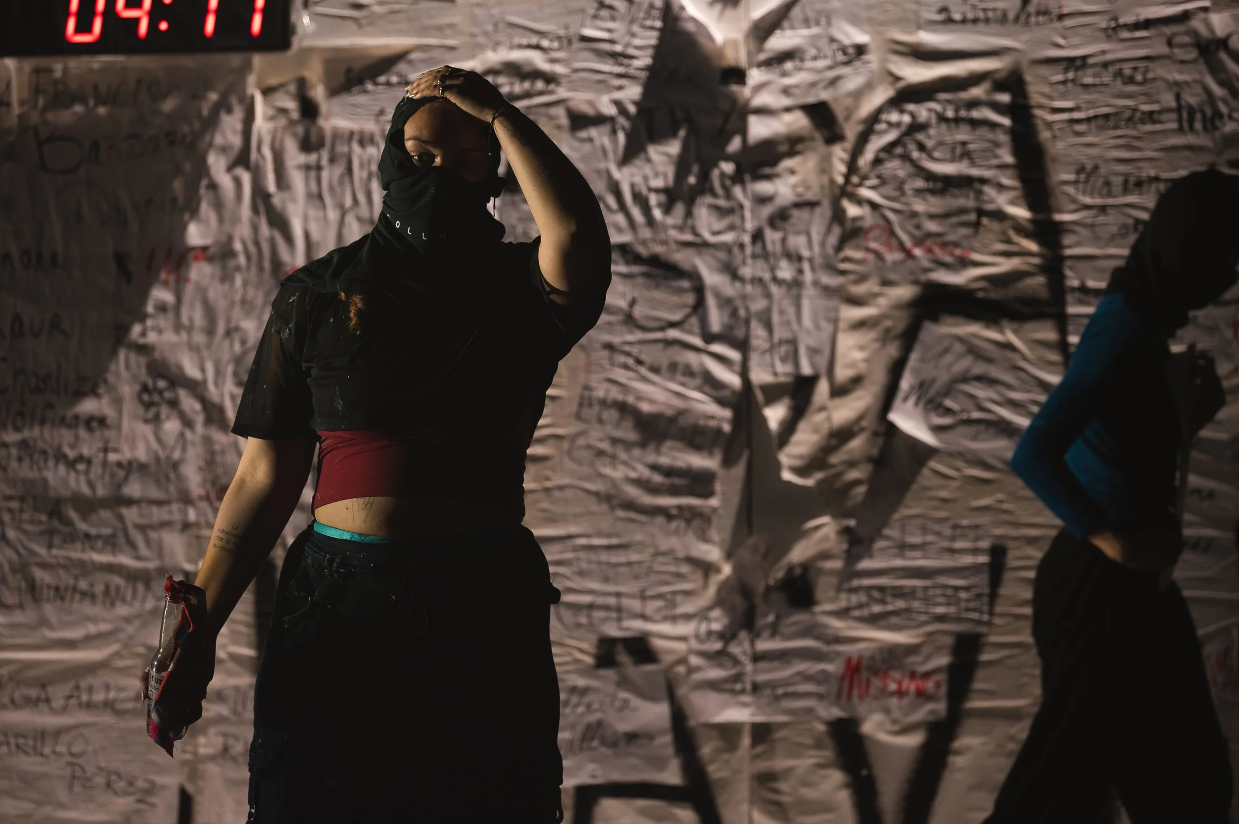 Une femme portant un foulard noir et un t-shirt noir se tient devant un mur recouvert de papiers ou de tissus écrits ou dessinés, dans un endroit sombre. Légèrement en arrière-plan, une autre personne avec une veste bleue et noire marche.