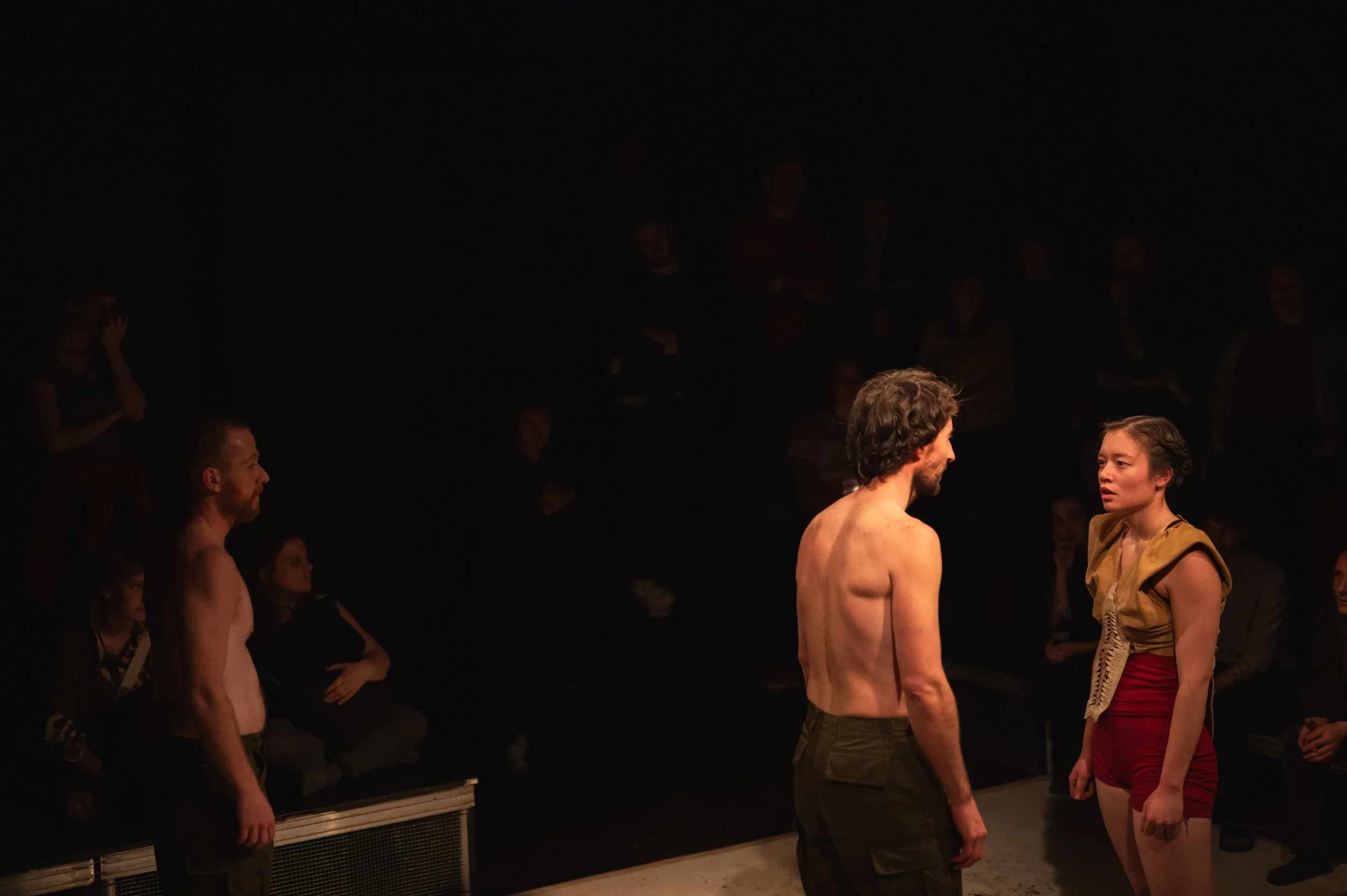 Deux acteurs nus face à face sur scène, un homme et une femme, avec un public en arrière-plan en arrière-plan dans une salle de théâtre sombre.