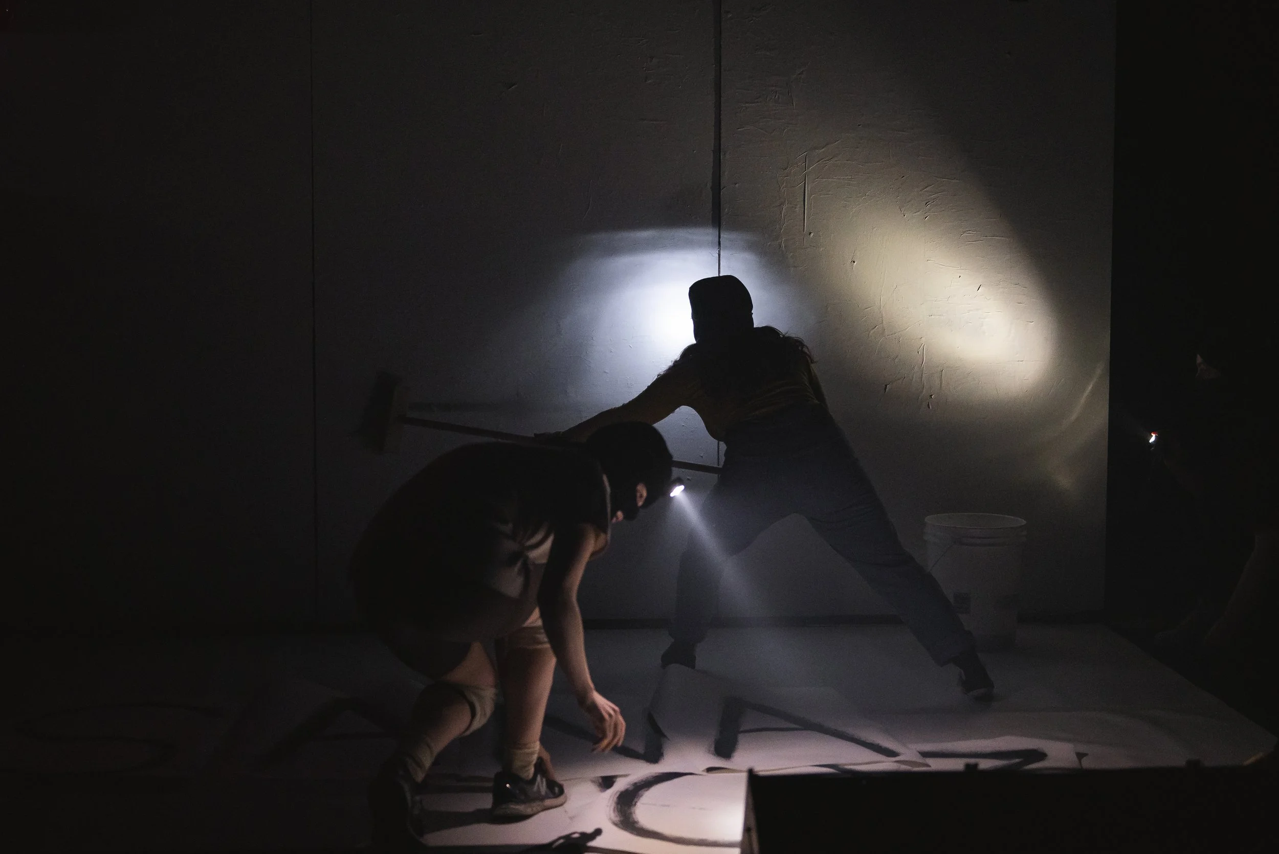 Deux personnes en silhouette dans une pièce sombre, utilisant des lampes de poche pour travailler. L'une d'elles, une femme, tient un balai ou une longue baguette, l'autre, un homme, est plié en dessous, tous deux concentrés sur une tâche au sol.