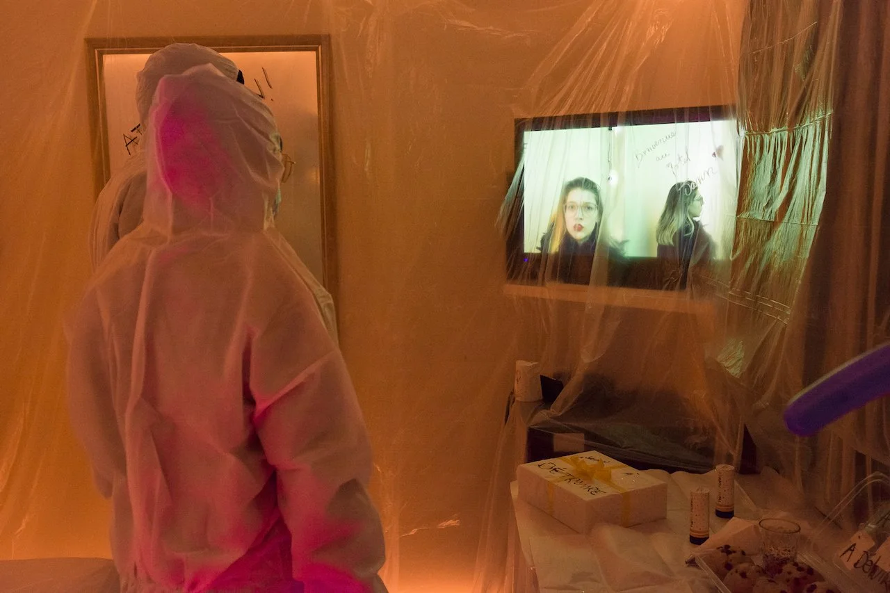 Deux personnes en tenue de protection regardent un écran avec une femme qui parle, dans une pièce éclairée par une lumière douce, avec des objets sur une table.