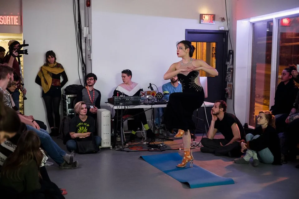 Une femme en robe noire et chaussures à talons oranges avec motifs se produit en tant que danseuse dans un spectacle de danse, entourée d'un public assis et debout dans une salle