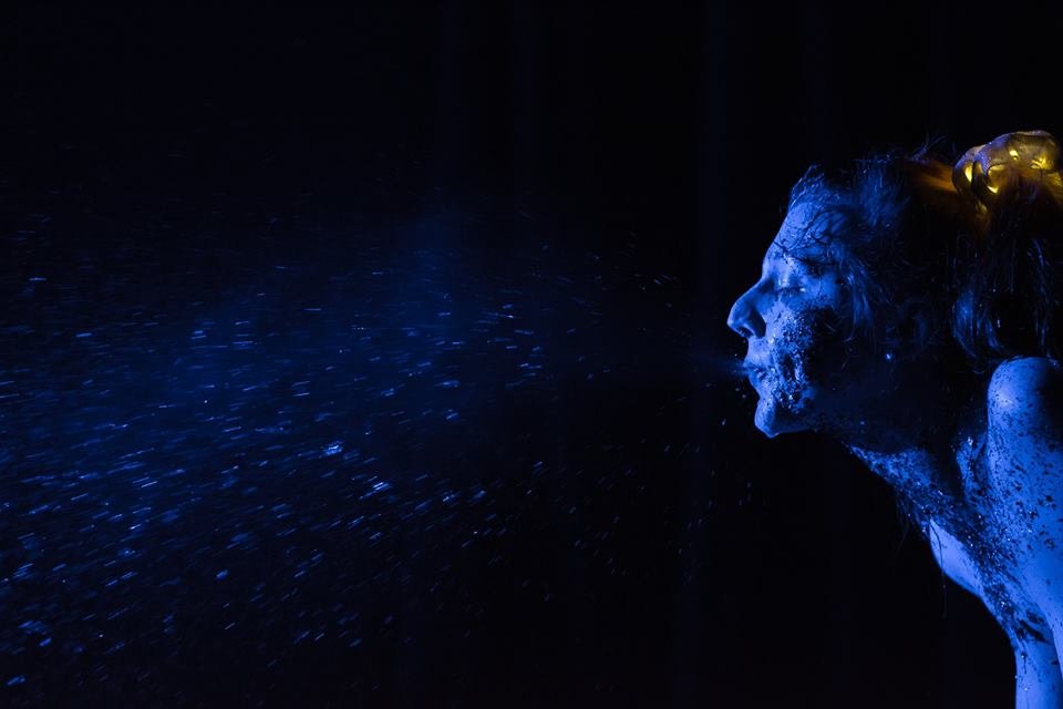 Une personne avec du maquillage ou peinture bleue, projetant de l'eau ou un liquide lumineux dans l'obscurité, avec des éclats de lumière bleue autour.
