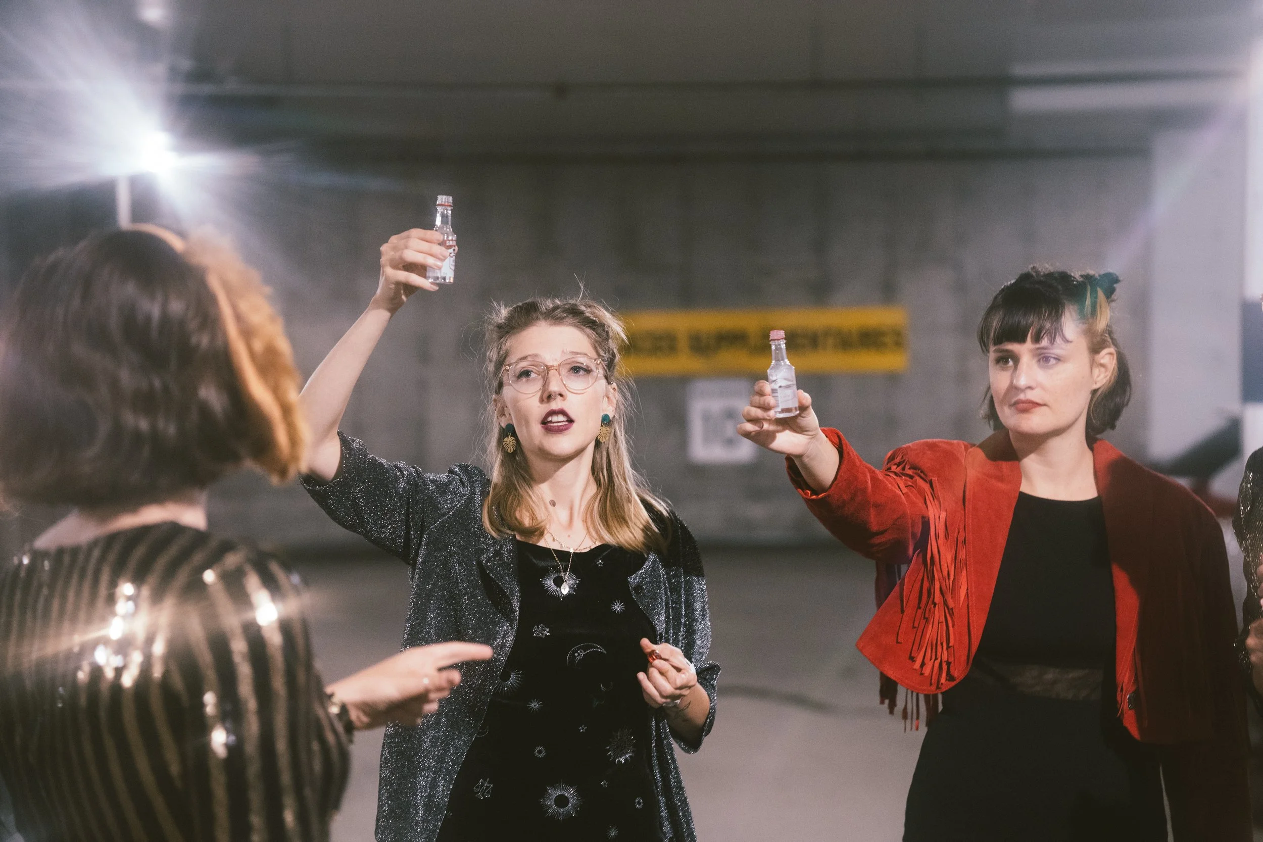 Trois femmes debout dans un parking, portant des vêtements élégants, tenant des bouteilles, semble être un toast ou une célébration.