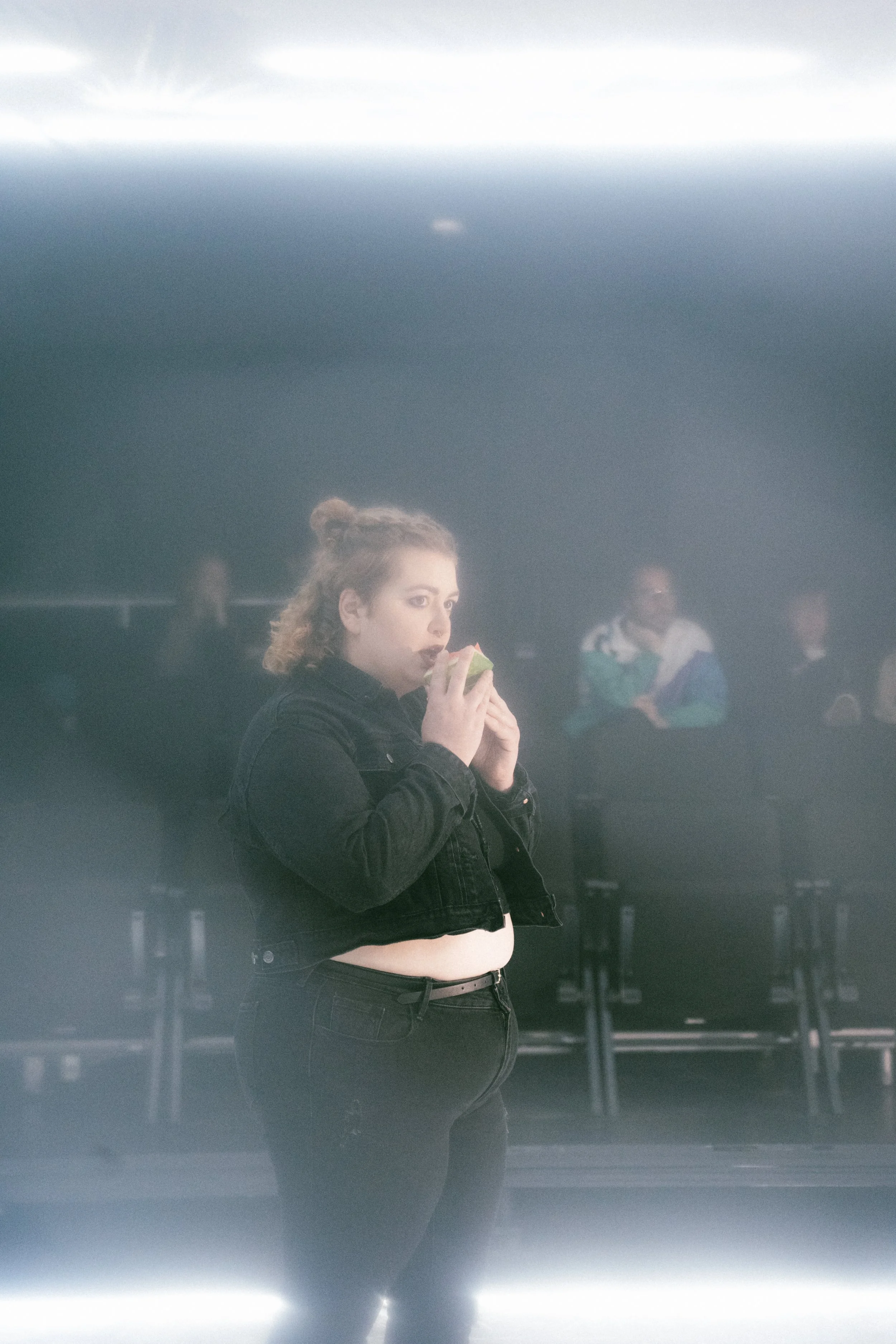 Une jeune femme avec une coiffure en chignon, portant une veste noire et un jean, mangeant une pomme dans un lieu sombre avec des personnes assises en arrière-plan.