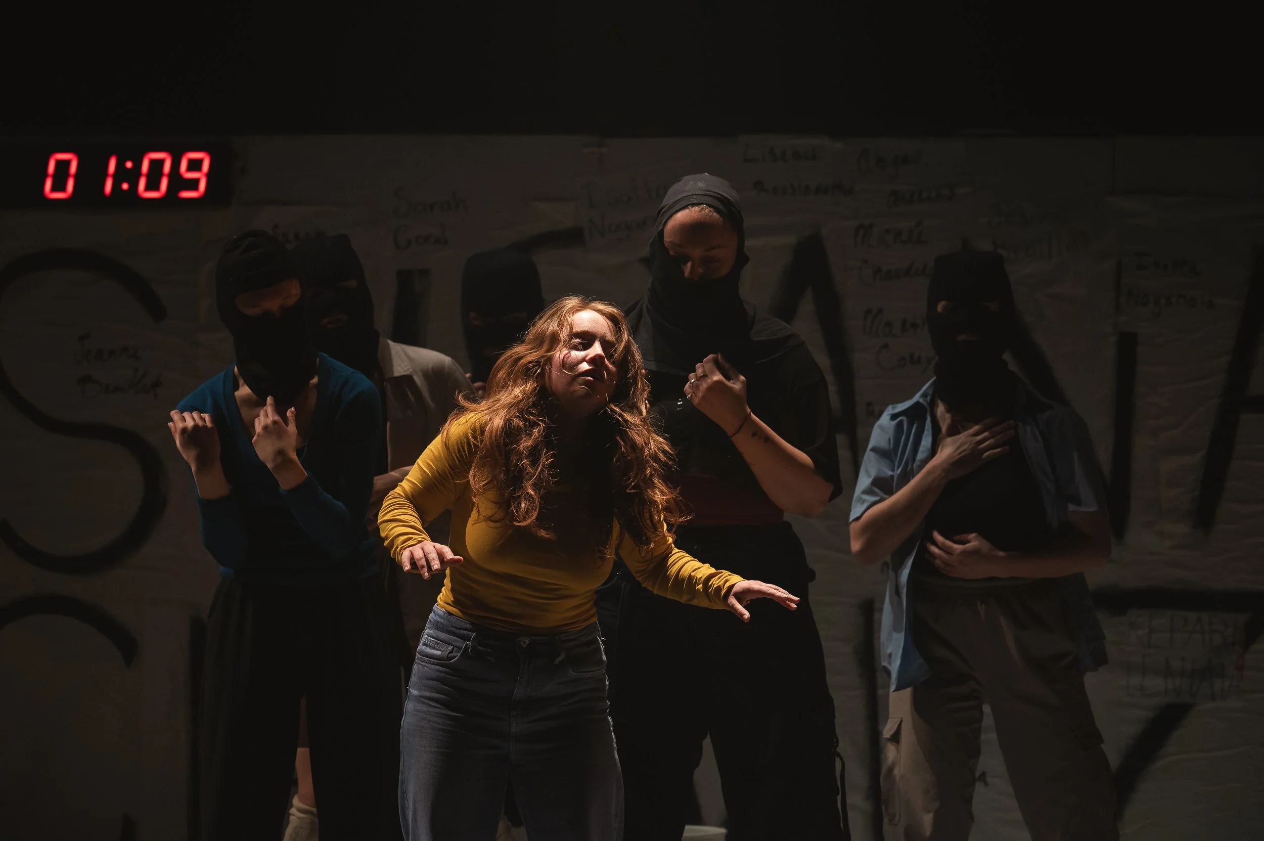 Une scène de théâtre avec plusieurs actrices sur scène, certaines portant des masques noirs, une femme en vêtements colorés au centre semble jouer un rôle intense. 