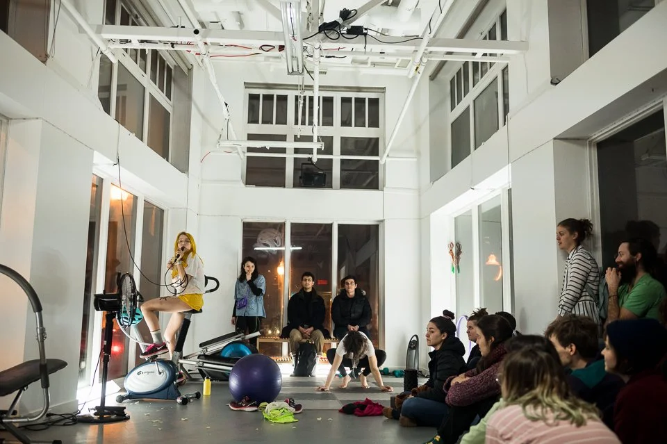Une femme avec un micro, assise sur un vélo d'appartement, parle à un groupe de personnes dans une salle lumineuse avec de grandes fenêtres.