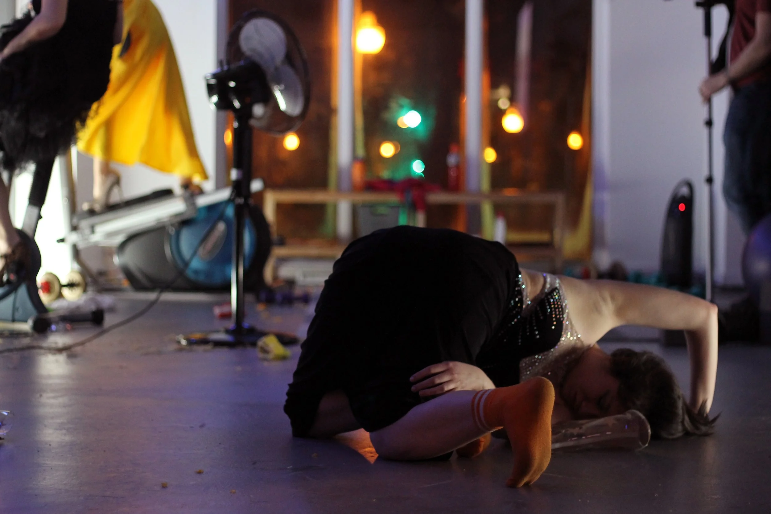 Une femme en costume scintillant est à terre, pliée en position de fœtus, dans une scène de spectacle ou de répétition avec des éclairages colorés en arrière-plan et des équipements de studio autour.