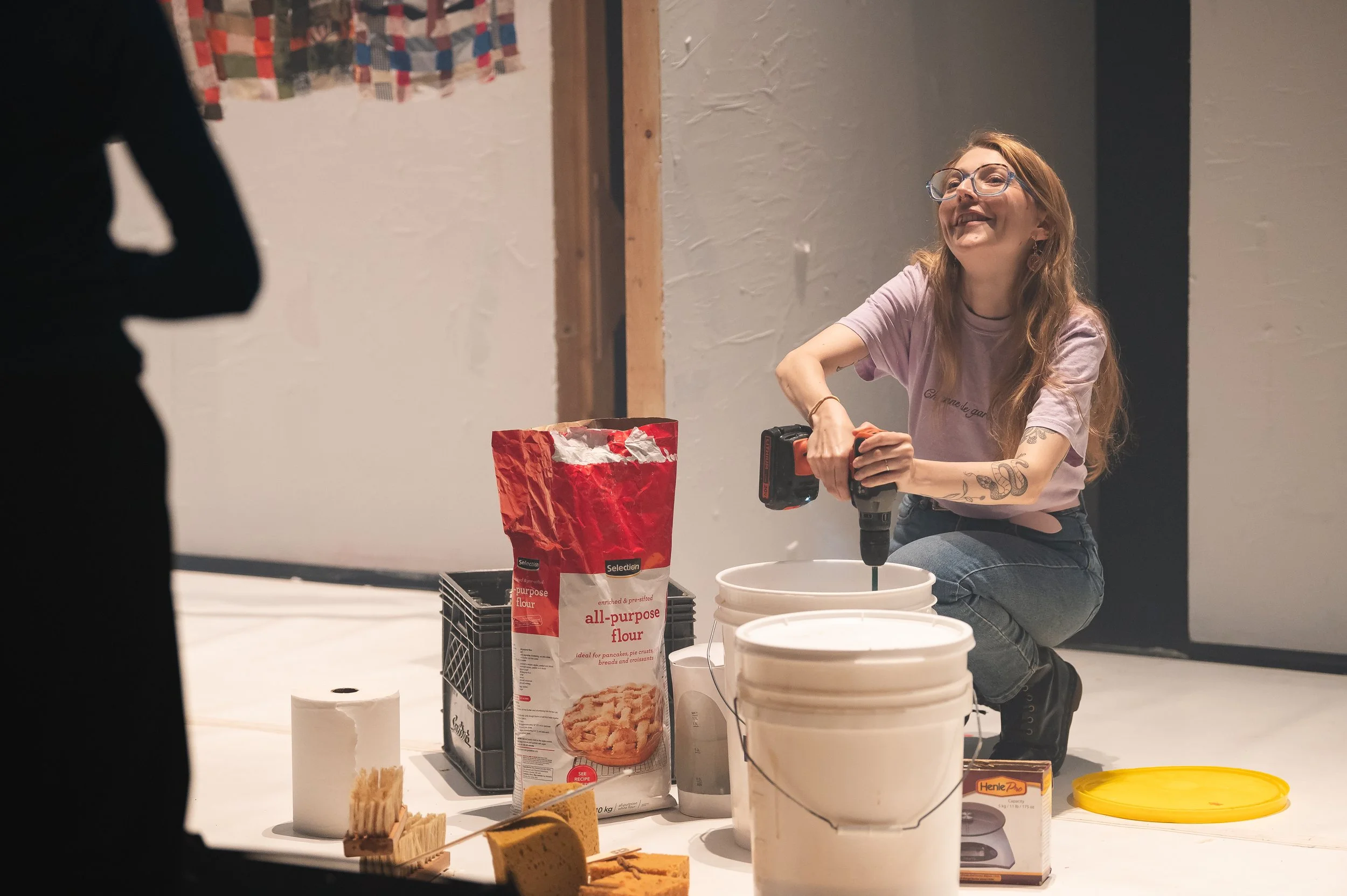 Une femme, portant des lunettes et un t-shirt violet, utilise une perceuse électrique sur une surface blanche, entourée de divers matériaux de construction et de maison comme du ruban adhésif, du papier toilette, un sac de farine, des pièces de cake 