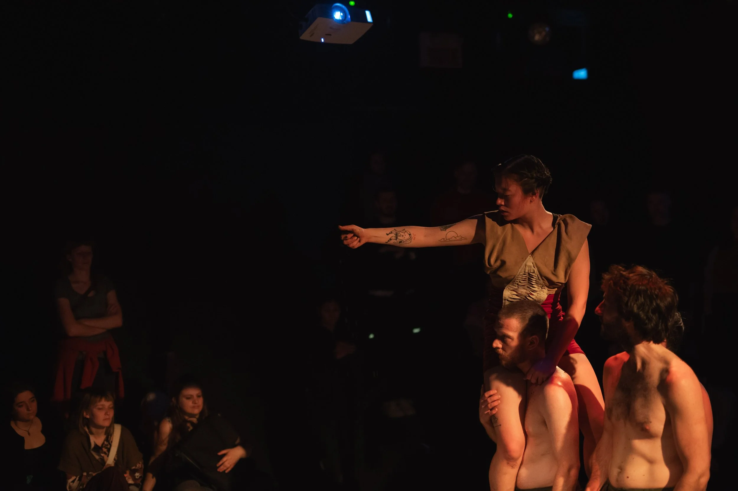 Une performance théâtrale ou artistique avec trois hommes nus portant une femme sur leurs épaules, dans un environnement sombre avec des spectateurs en arrière-plan.