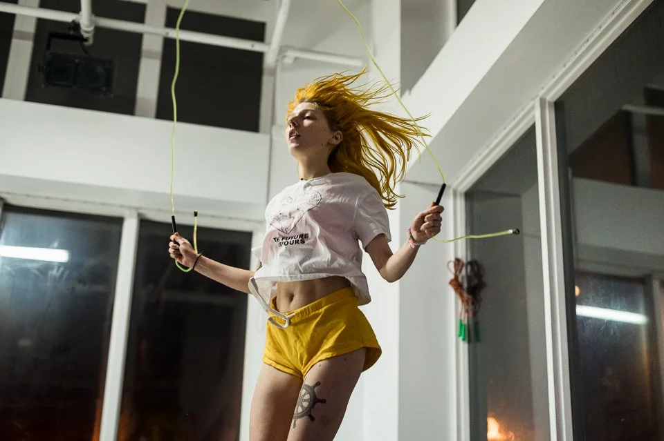 Une jeune femme aux cheveux rouges danse avec un saut à cordes à l'intérieur, portant un t-shirt blanc, des shorts jaunes, et un tatouage de soleil sur la cuisse. Elle semble s'amuser.