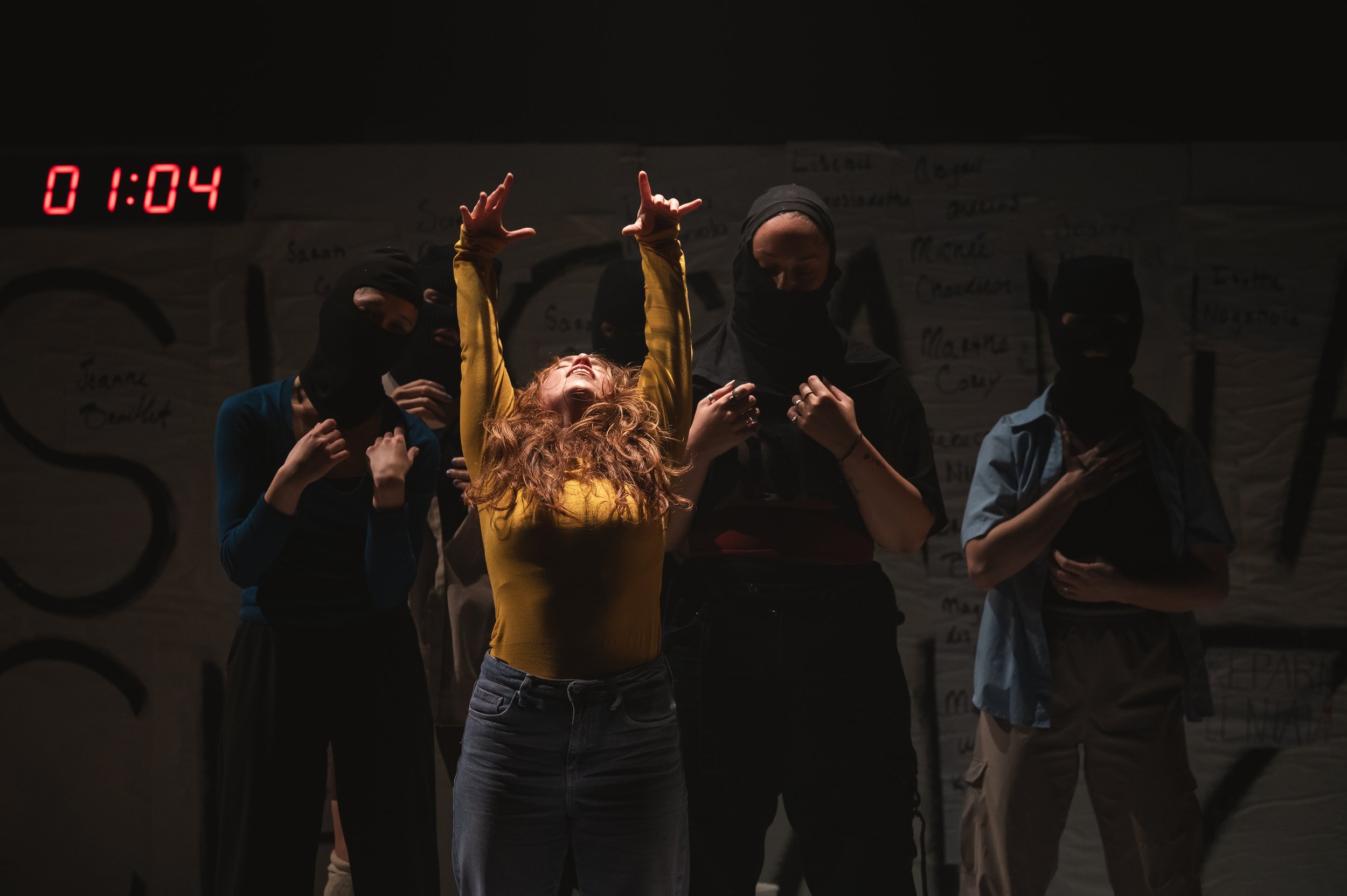 Une jeune femme avec des cheveux bouclés roux, vêtue d'un pull jaune et pantalon en jean, lève les bras, les mains en gesture expressive, en pleine performance dramatique sur scène, entourée de personnes portant des masques noirs et des vêtements sombres, sous un éclairage tamisé, avec un chronomètre orange au coin supérieur gauche affichant 01:04.