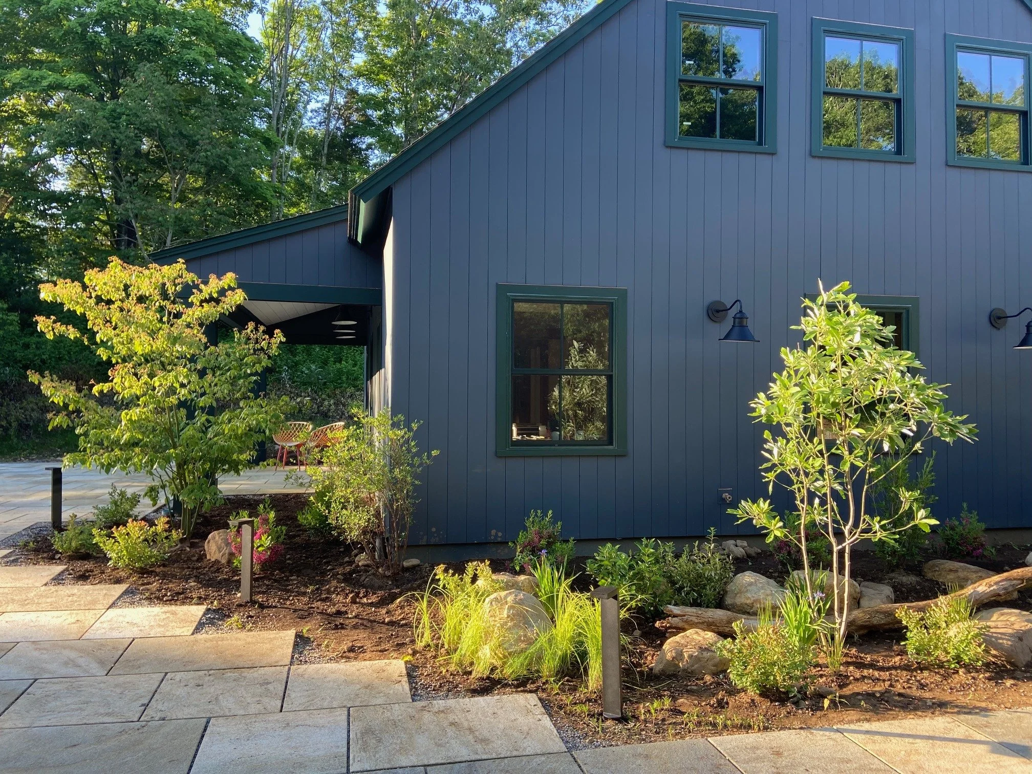 A modern house with blue vertical siding and dark green trim, surrounded by landscaped garden with small trees, bushes, and rocks, and a stone pathway.