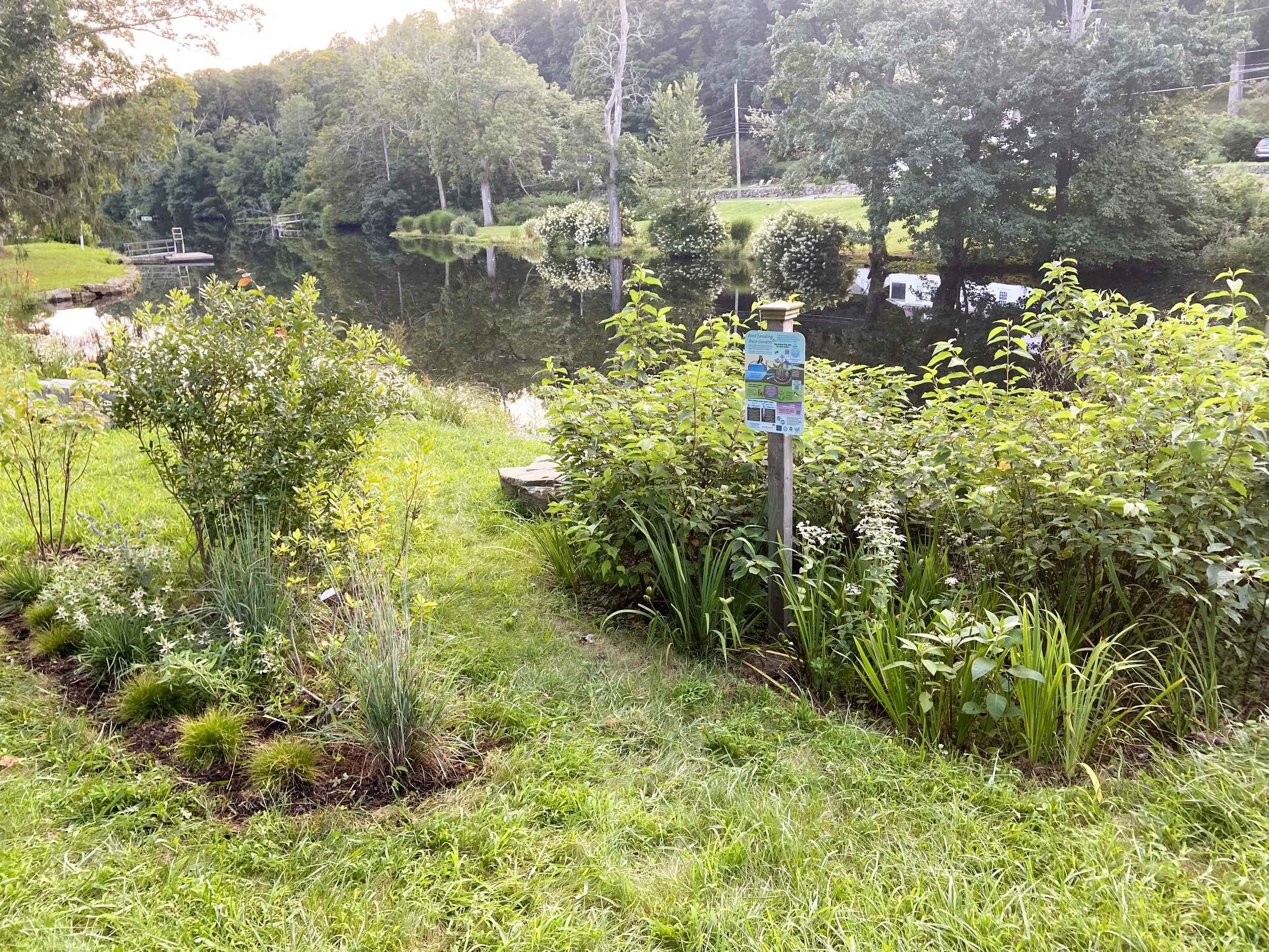 A grass and flower garden next to a calm river with trees, bushes, and a small dock reflected in the water.