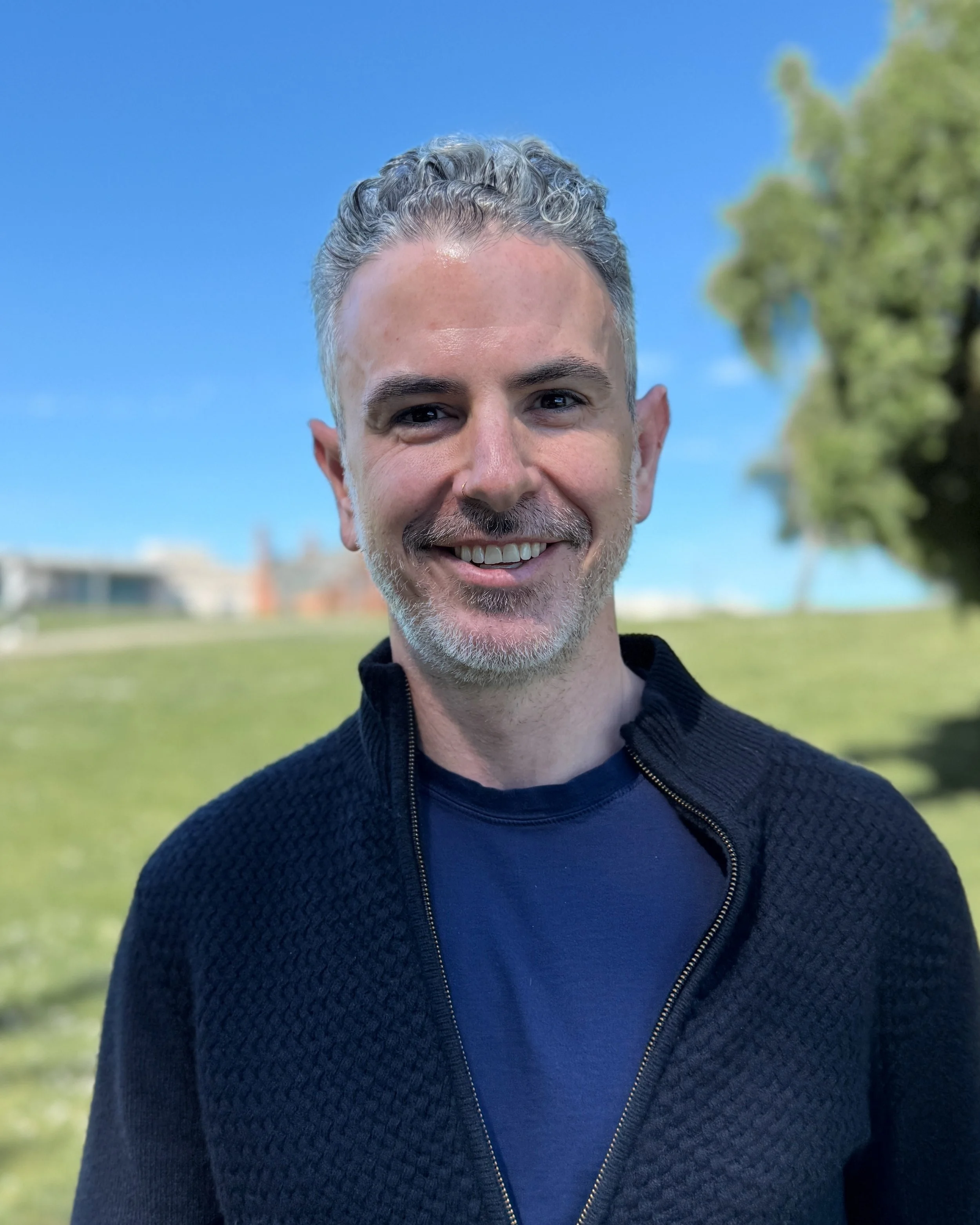 A man with short curly gray hair and a beard smiling outdoors, with a blue sky and green trees in the background.