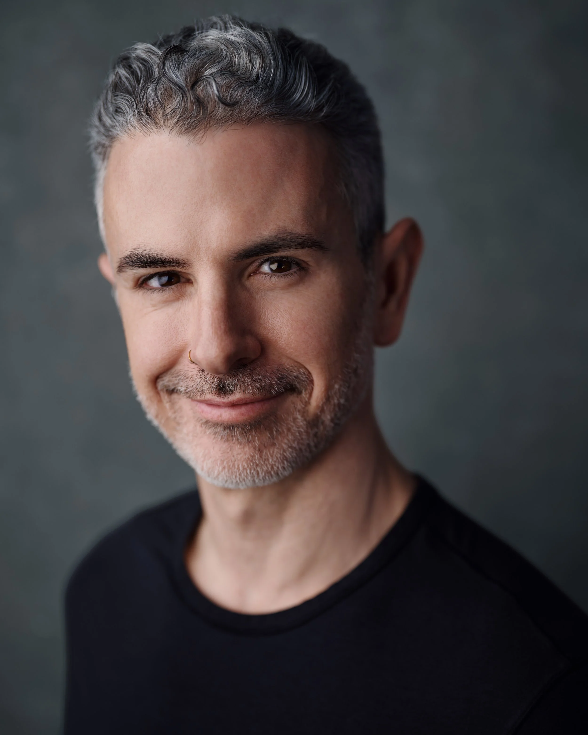 A close-up portrait of a smiling middle-aged man with salt-and-pepper hair, a light beard, and mustache, wearing a black shirt against a dark background.