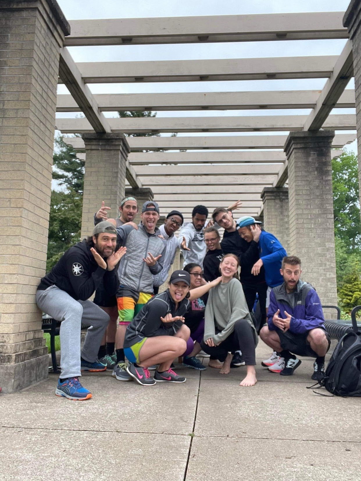 Group of people posing and smiling outdoors under a pergola on a cloudy day, with trees in the background.