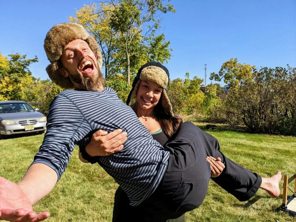 A man dressed in a striped shirt and furry winter hat being carried by a woman in a black jacket and furry winter hat outdoors on a sunny day with trees and parked cars in the background.