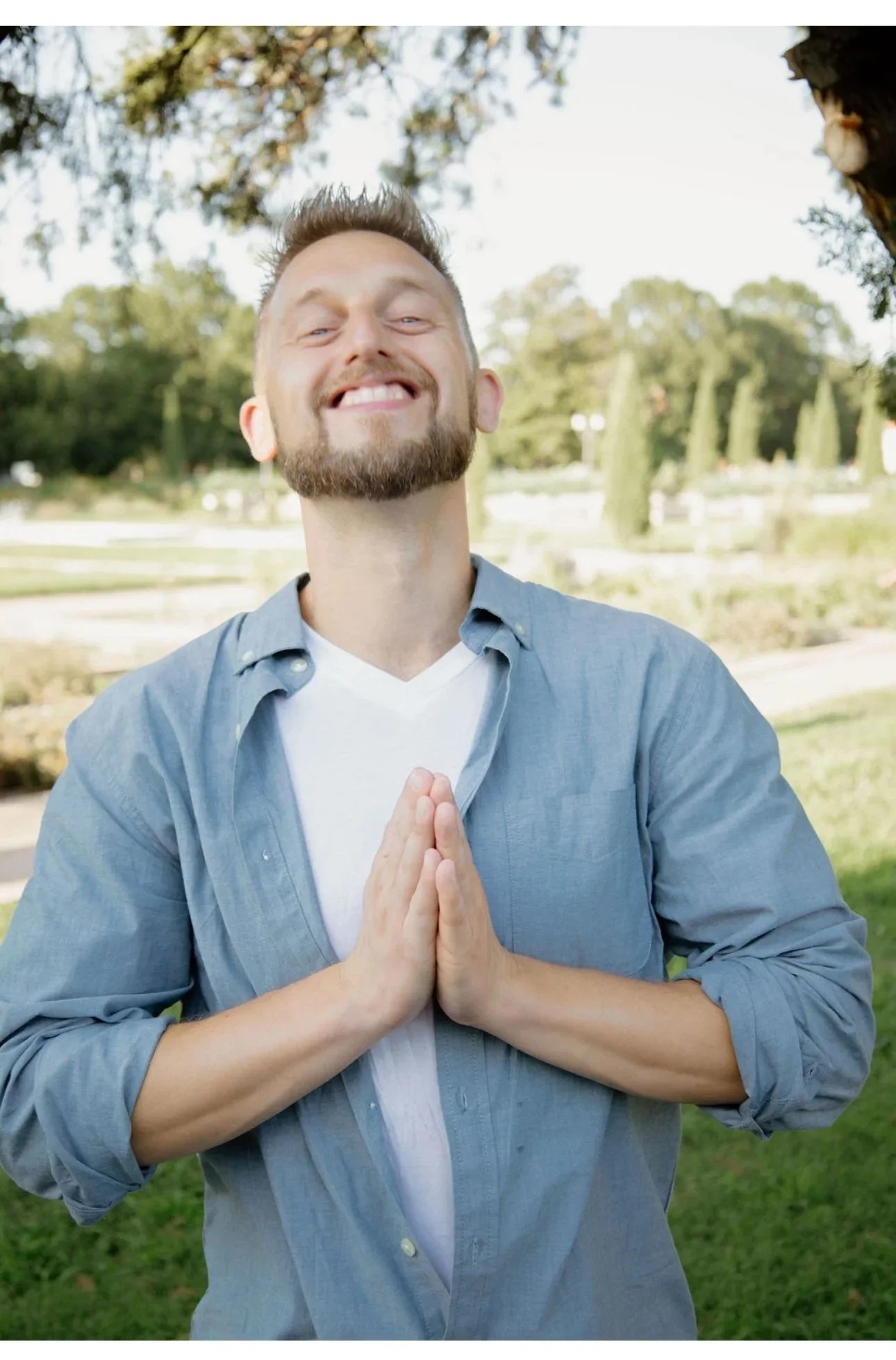 A smiling man with a beard and mustache standing outdoors in a park, with his hands pressed together in a prayer-like gesture.