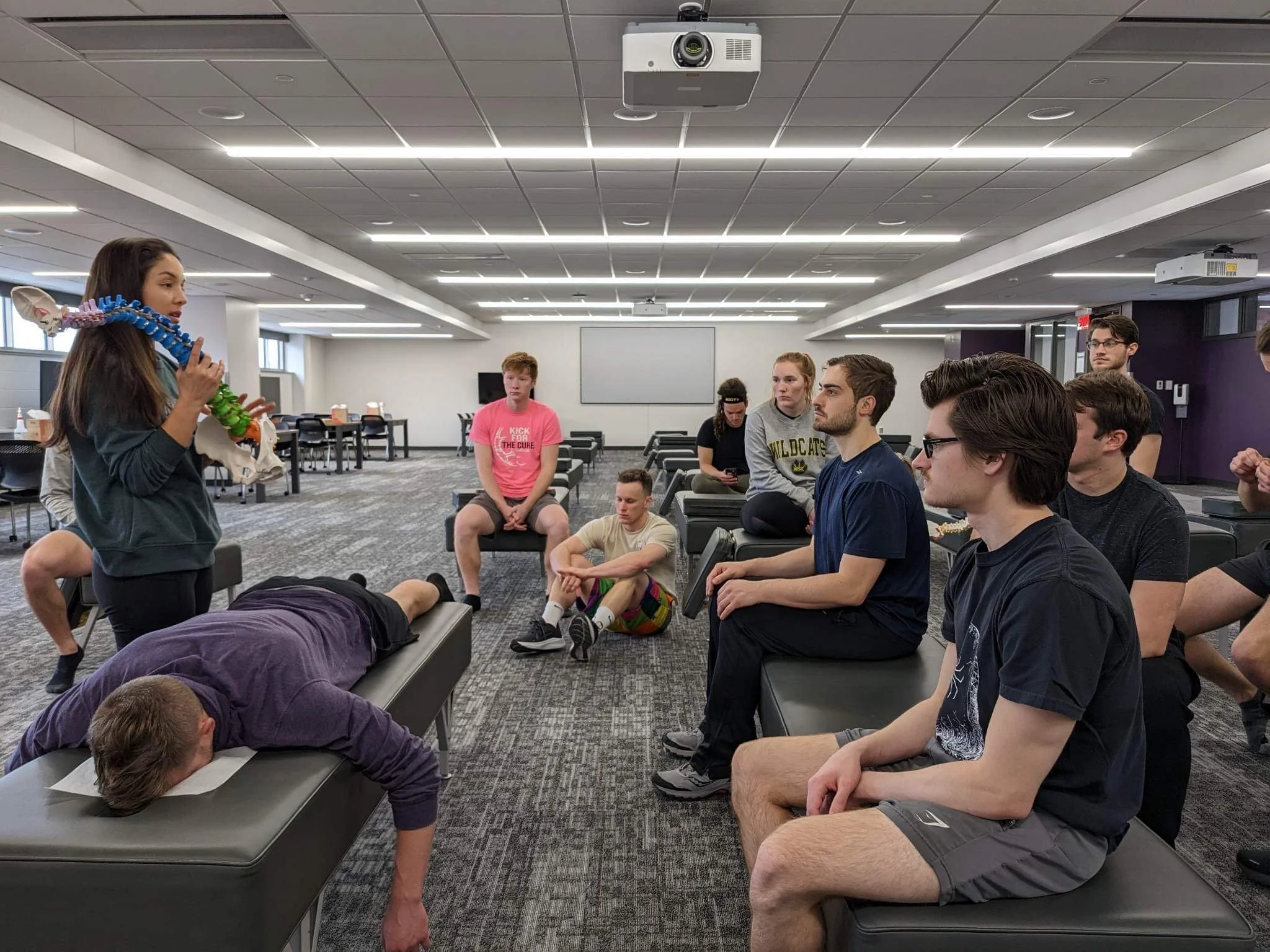 A woman holding a model of a human skeleton demonstrates to a group of young adults seated in a classroom or conference room.