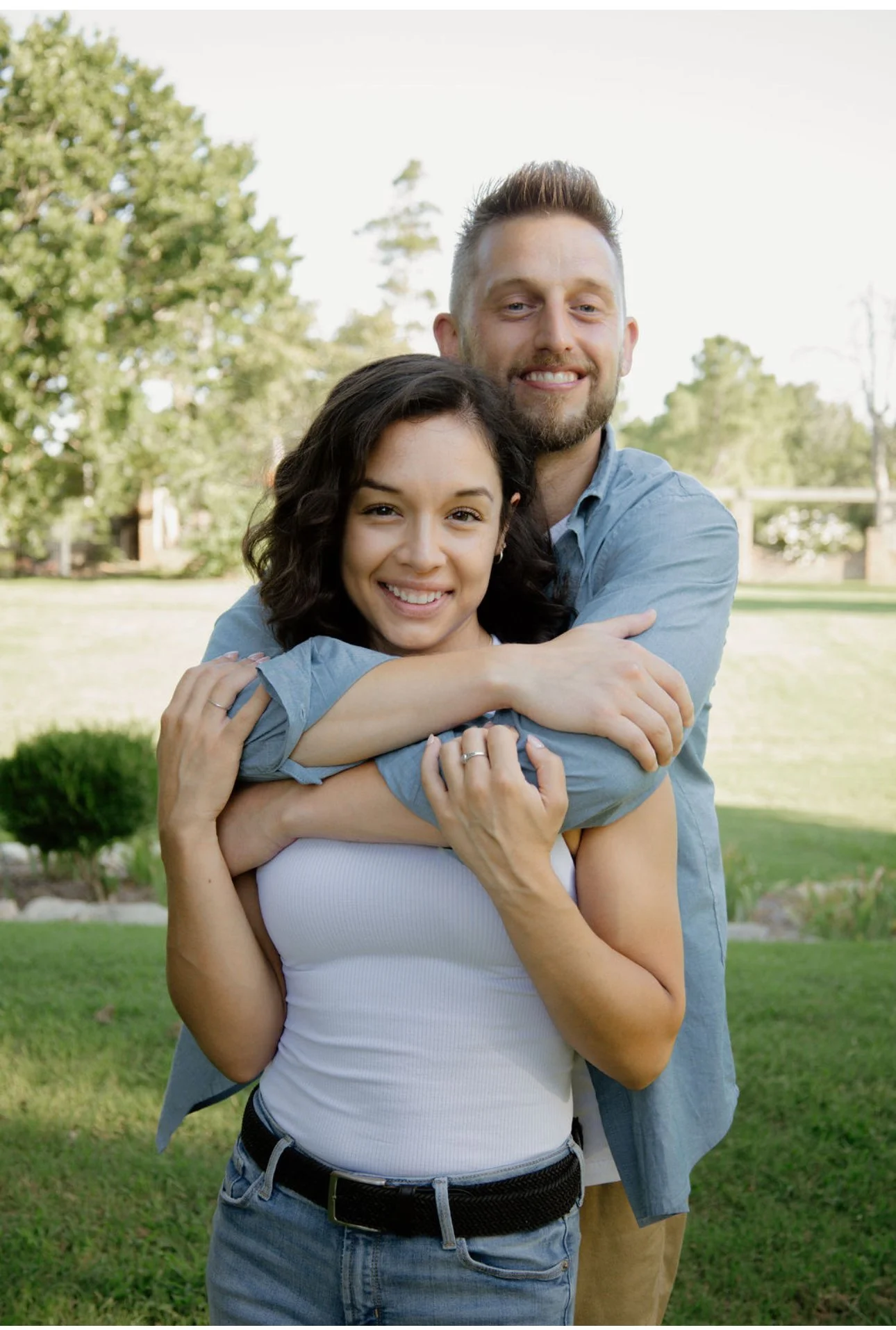 A happy couple hugging outdoors in a park with trees and grass in the background.