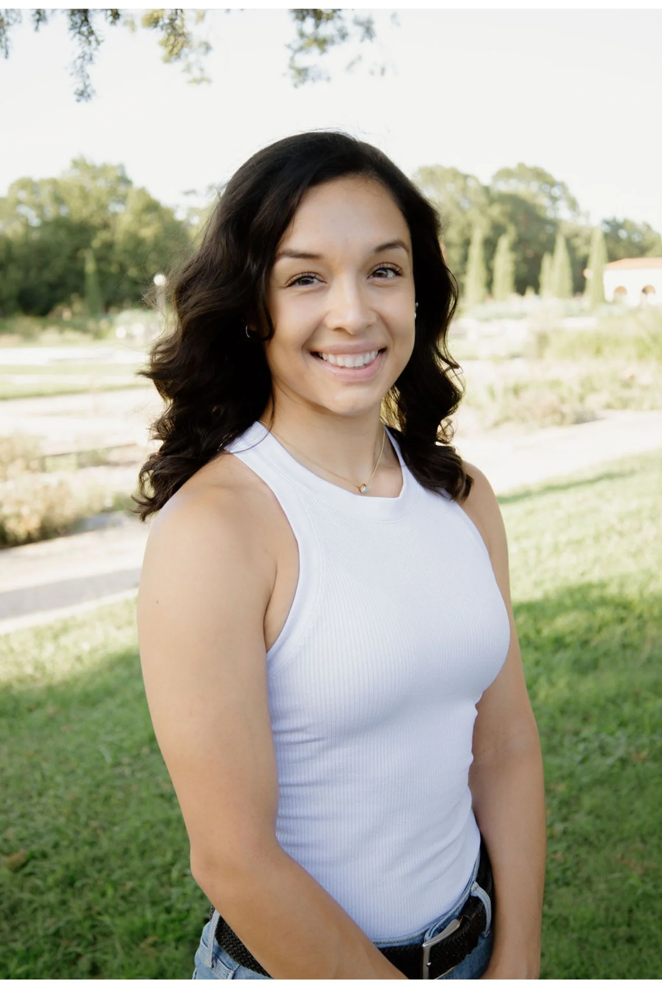 A smiling young woman with dark, wavy hair in a sleeveless white shirt standing outdoors in a park with green grass, trees, and a cloudy sky in the background.