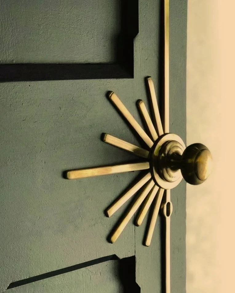 Close-up of a modern wall clock with a brass finish, showing Roman numerals and a minimalist design, mounted on a textured green wall.