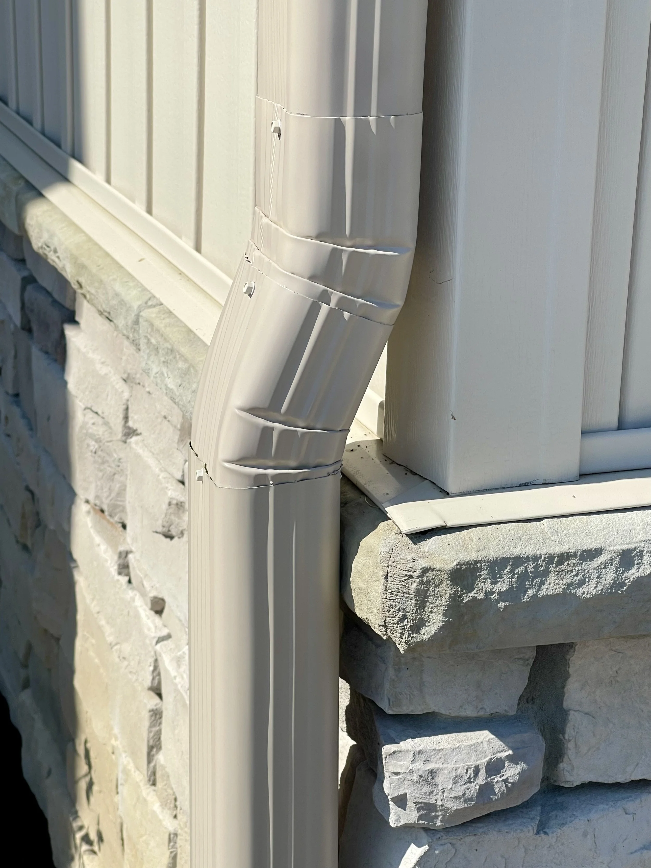 Outdoor close-up of a beige metal downspout attached to a house with stone foundation and beige siding.
