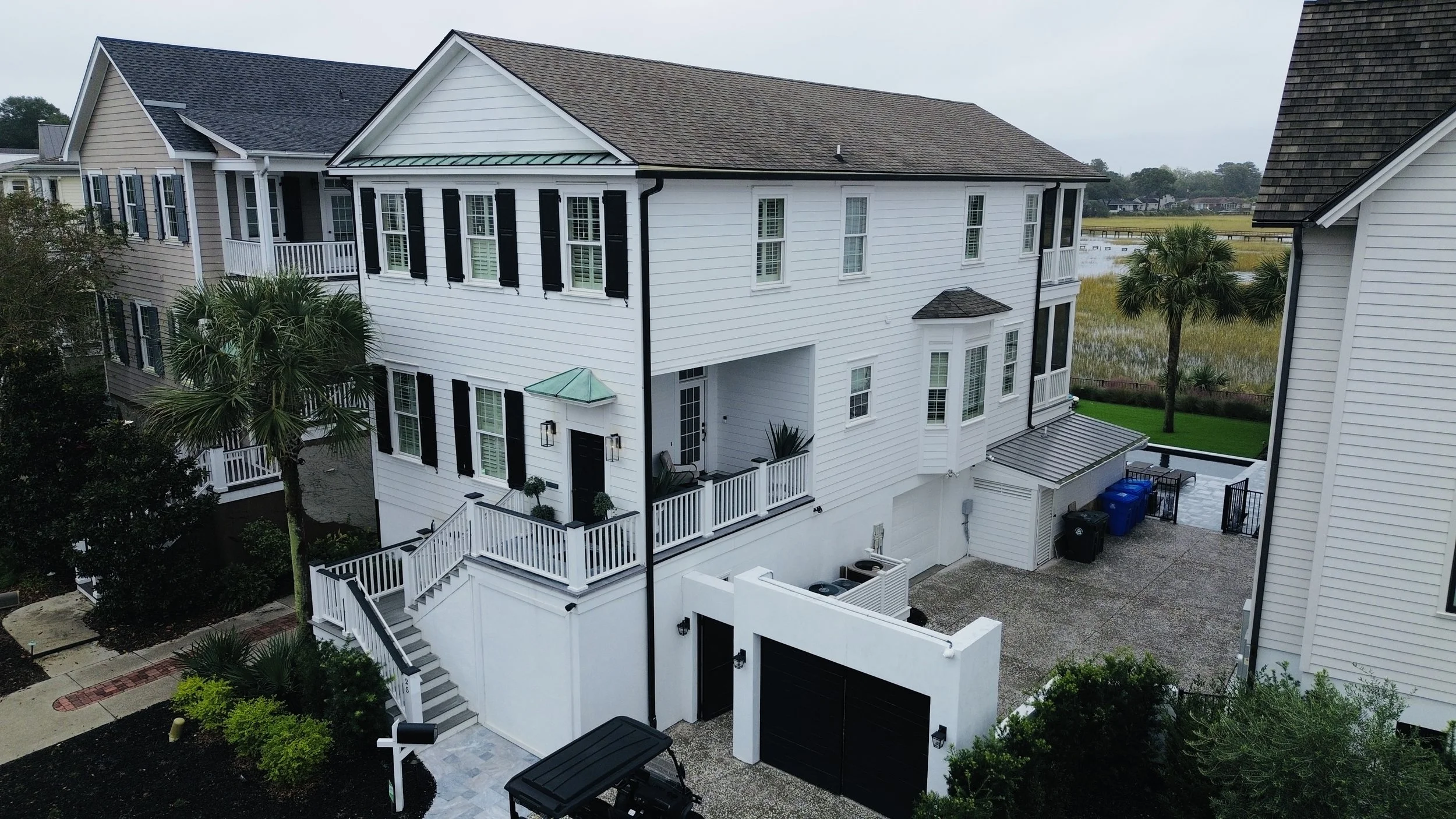 A white multi-story house with black shutters, a small front porch with stairs, a backyard with outdoor furniture, palm trees, and a water body in the background.