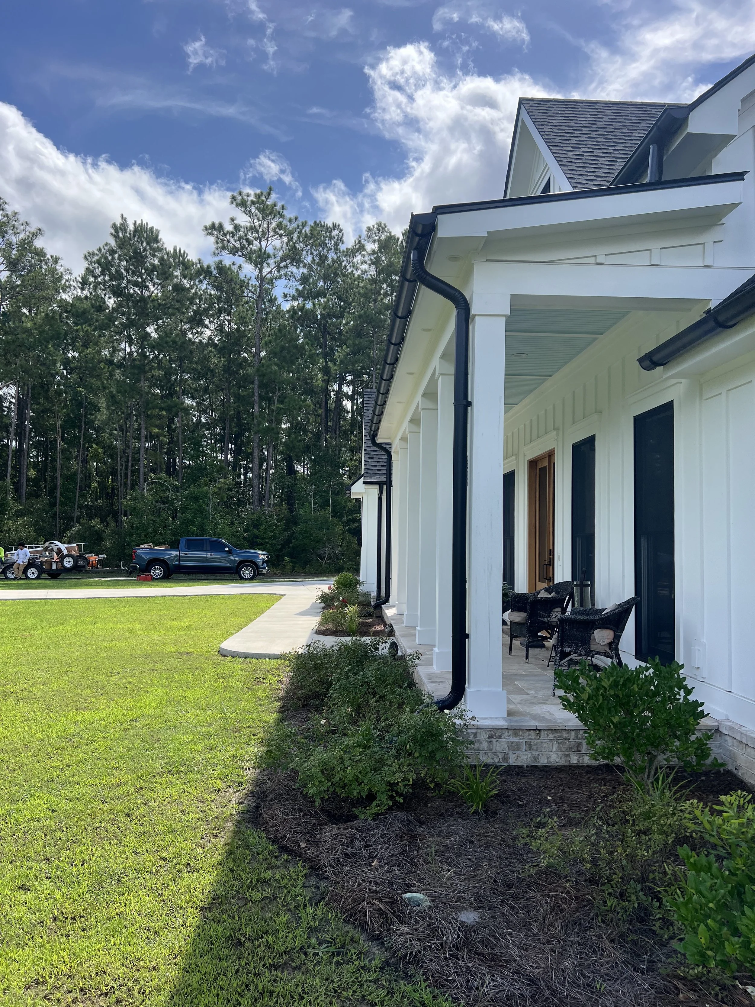 Front porch of a white house with black gutters and chairs, green lawn, driveway with a truck, and trees in the background under a blue sky with clouds.